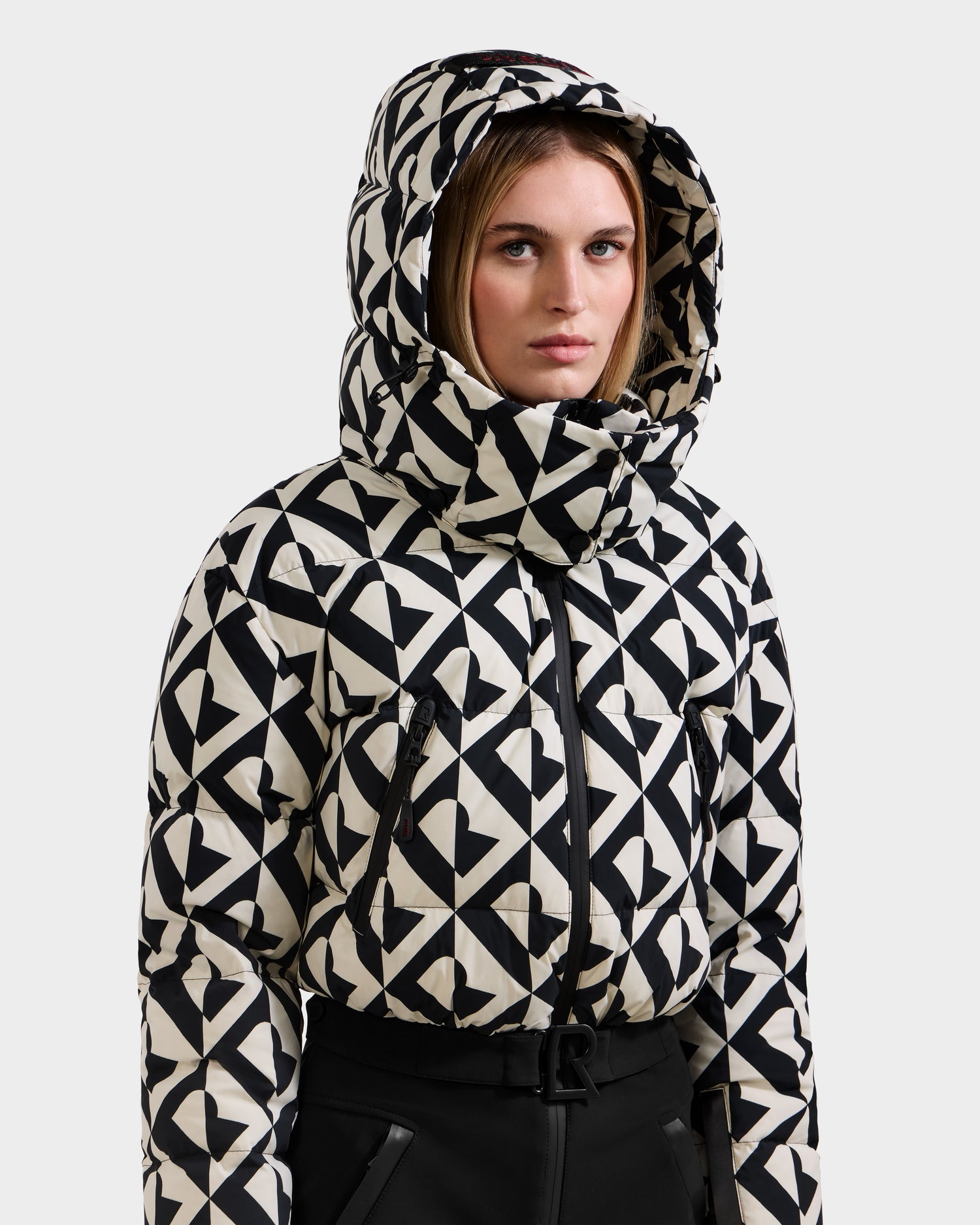 A woman models the Rudsak DINARA - 8125600 Vanilla Monogram hooded puffer jacket, featuring a bold geometric print, high collar, and zippered pockets, inspired by elements of a women's ski suit.