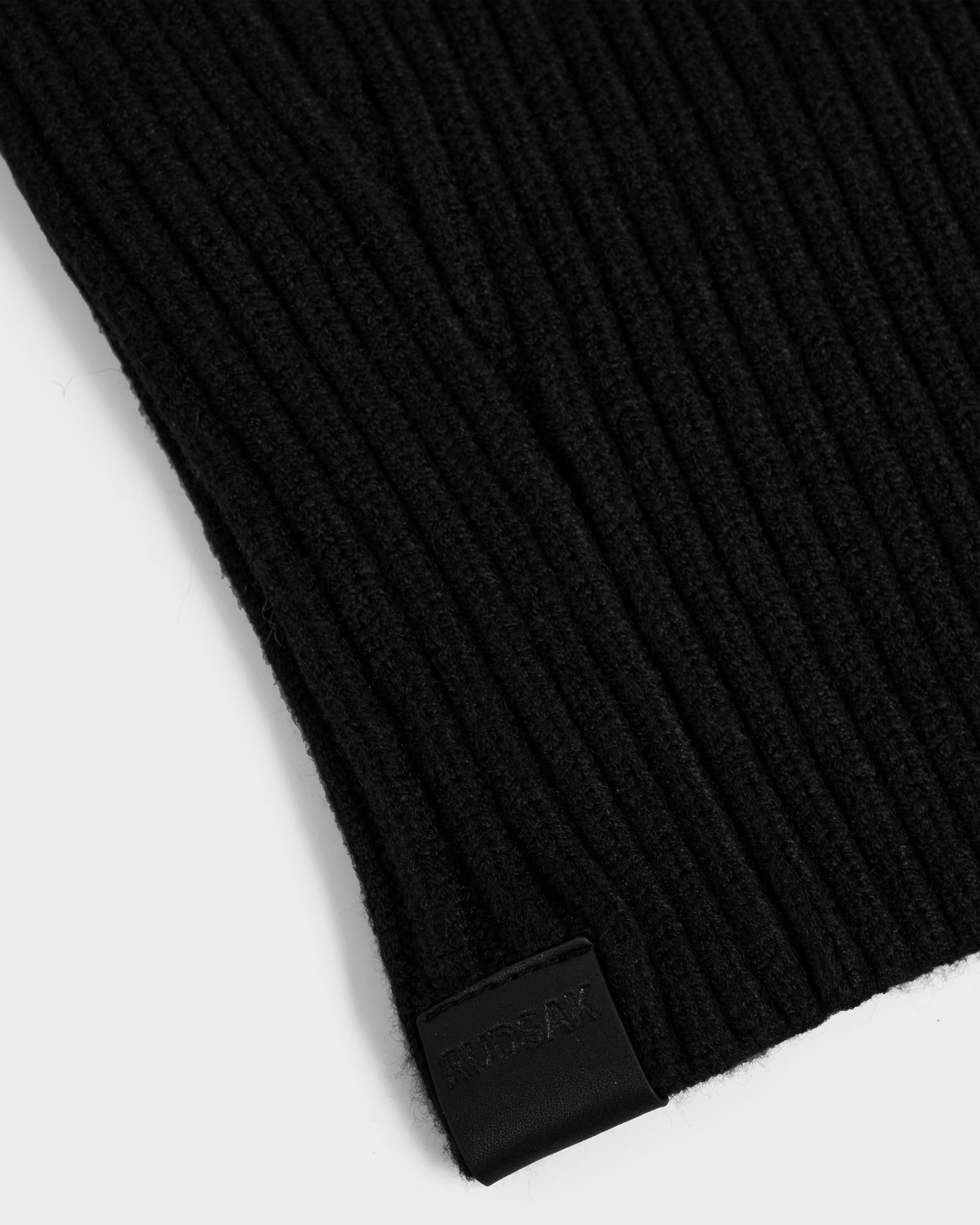 Close-up of the Rudsak DIXIE - 8425551 Black ribbed knit balaclava, featuring a small black embossed tag at the edge, set against a plain white background.