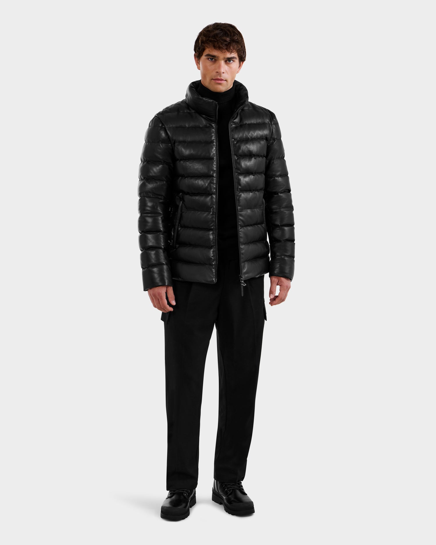A man stands facing forward, wearing the Rudsak DRAVEN X - 6125686 Black leather puffer over a black turtleneck, pants, and shoes, set against a plain light background.