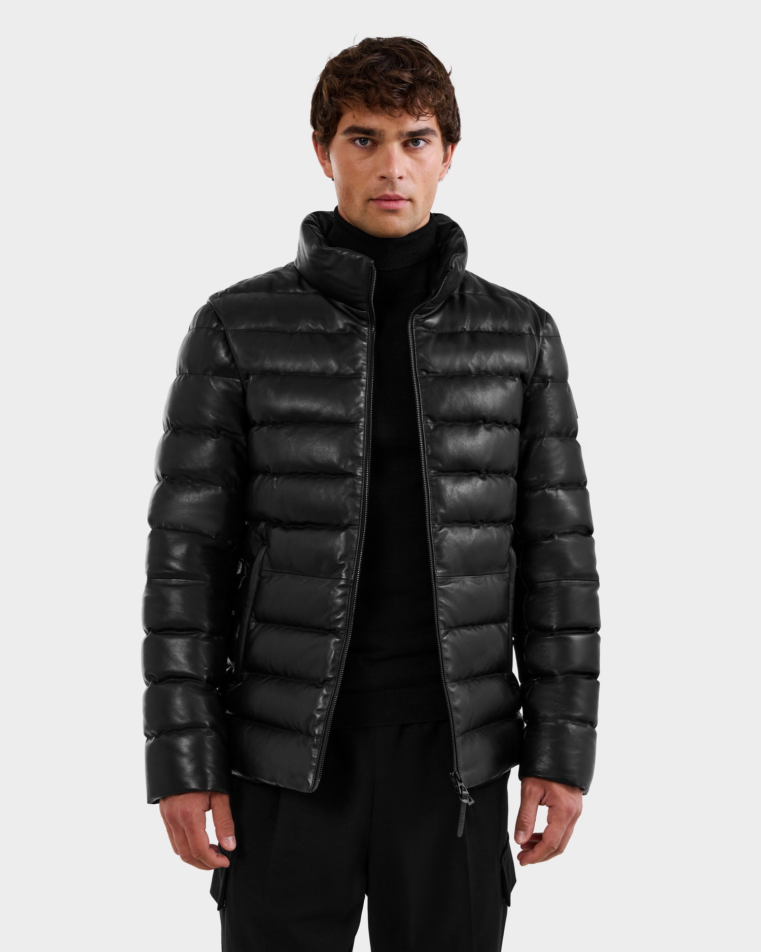 A young man stands facing forward, wearing the Rudsak DRAVEN X - 6125686 Black leather puffer jacket over a black turtleneck and black pants, set against a plain light gray background.