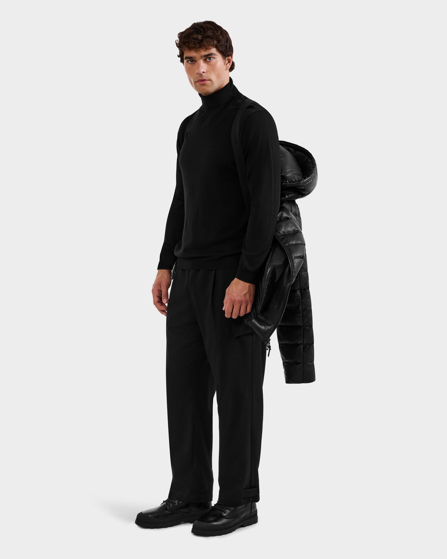 A man faces forward in a black turtleneck, pants, and shoes, holding the Rudsak DRAVEN X - 6125686 Black men’s leather down jacket over his left shoulder against a plain light gray background.