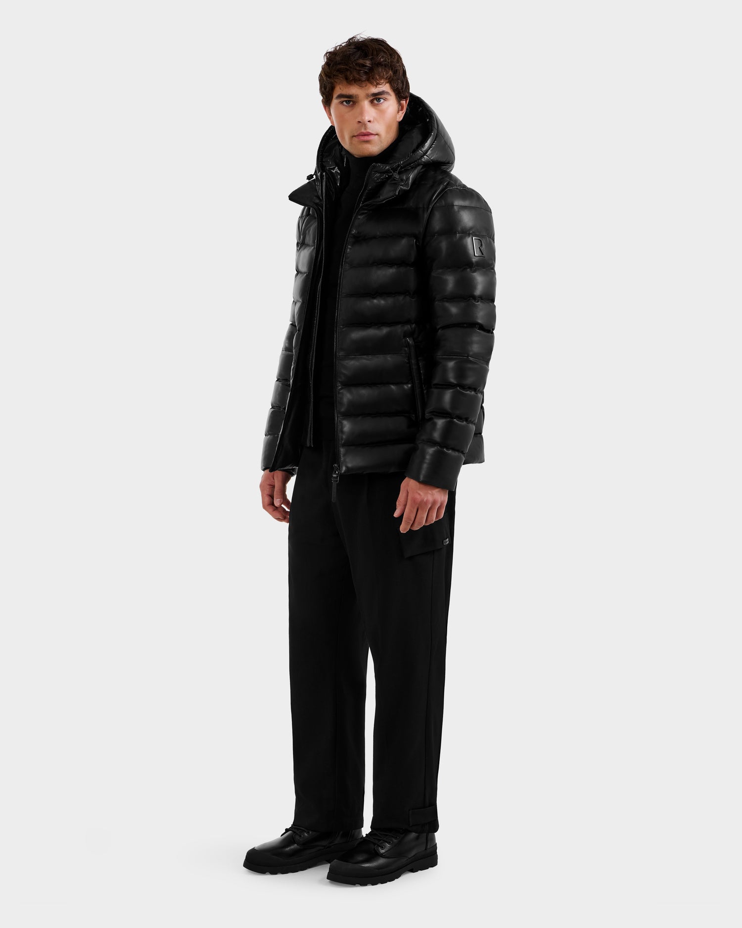 A man stands against a plain background wearing the Rudsak DRAVEN X - 6125686 Black men’s leather down jacket with a hood, paired with black pants and shoes. He faces forward neutrally, hands relaxed at his sides.