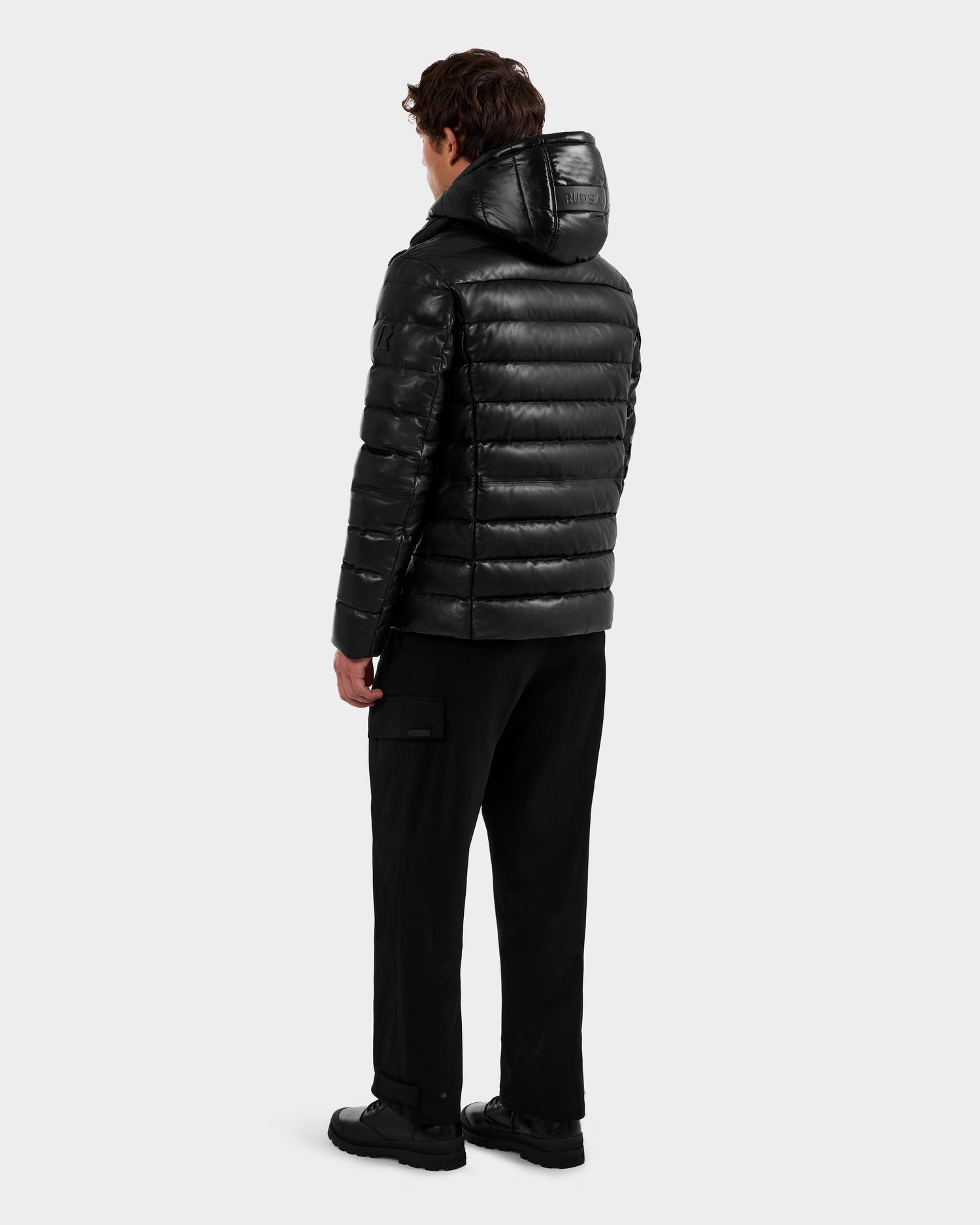 A person faces away, wearing the Rudsak DRAVEN X - 6125686 Black leather puffer jacket with hood, black pants, and black shoes, set against a plain light gray background.