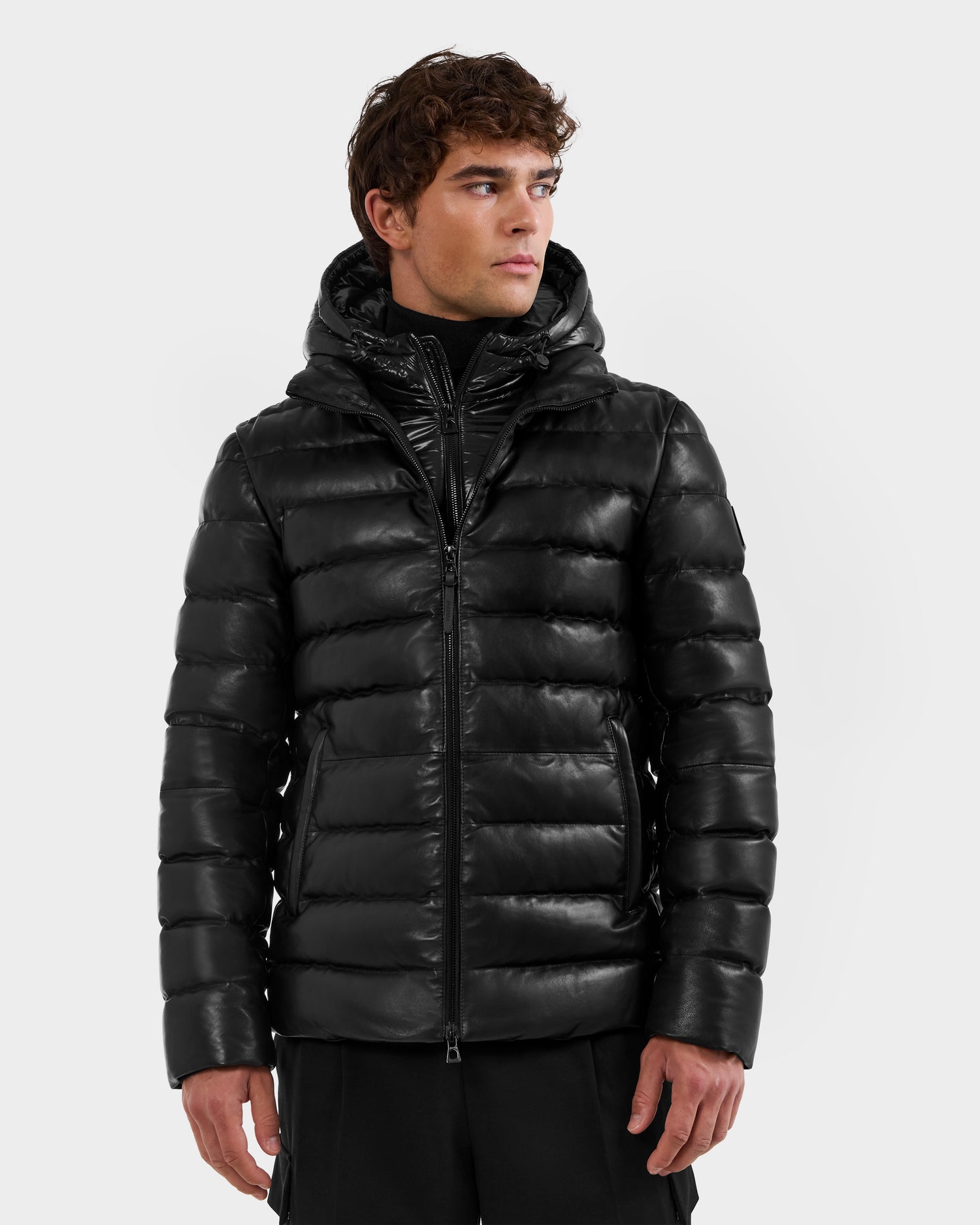 A man in a shiny black Rudsak DRAVEN X - 6125686 leather puffer jacket with hood stands against a light gray background, looking left with a neutral expression.
