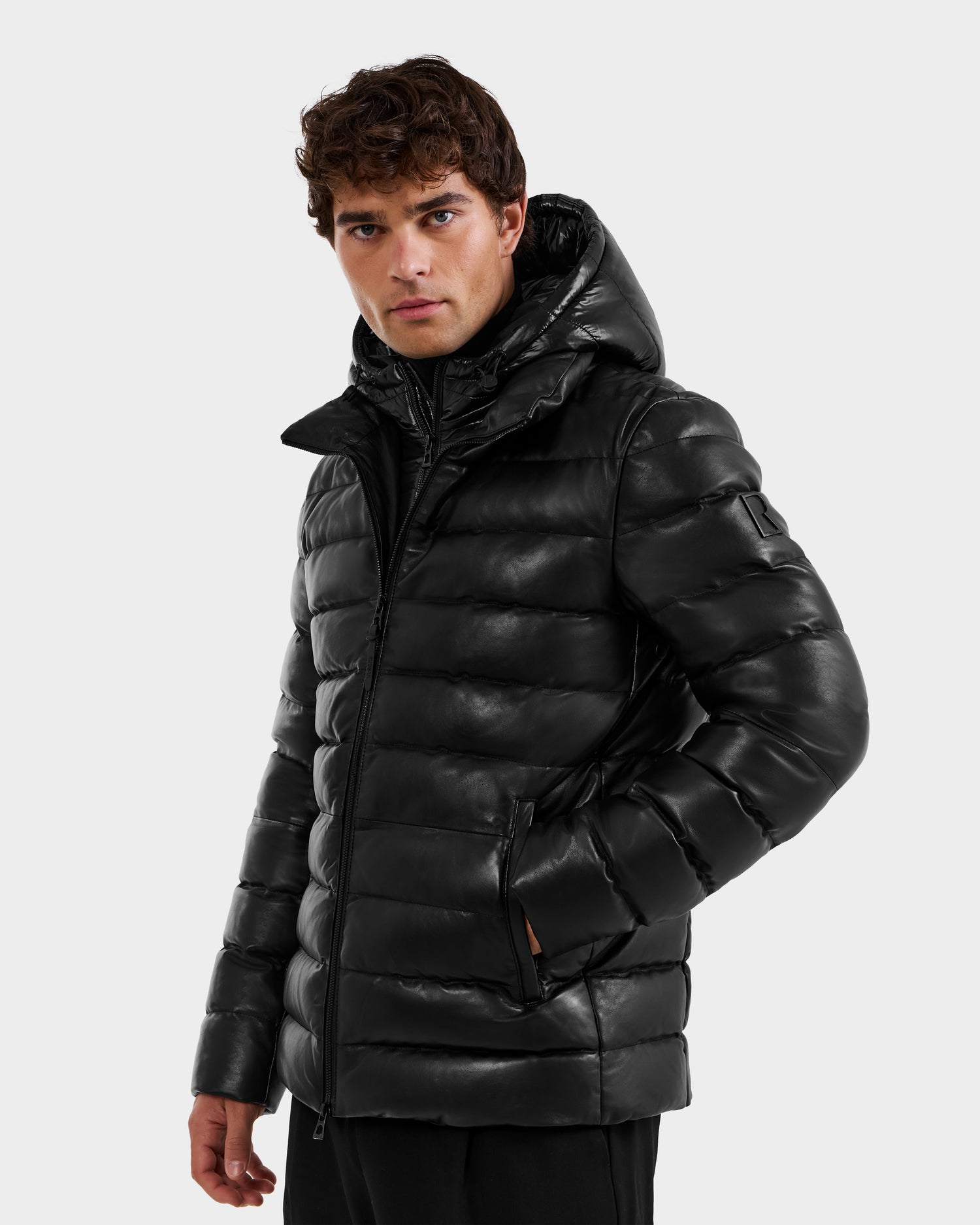 A man wearing the Rudsak DRAVEN X - 6125686 Black men’s leather down jacket with a hood stands against a plain light gray background, looking slightly to the side.