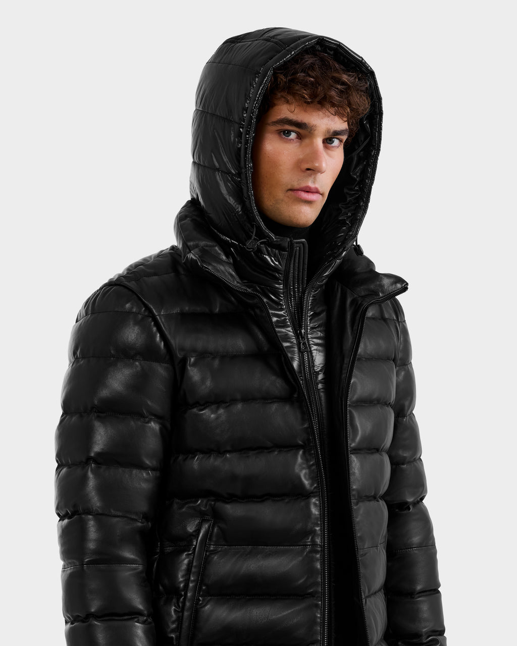 A young man with curly hair wears a shiny black Rudsak DRAVEN X - 6125686 men’s leather down jacket with the hood up, looking slightly to the side against a plain light background.