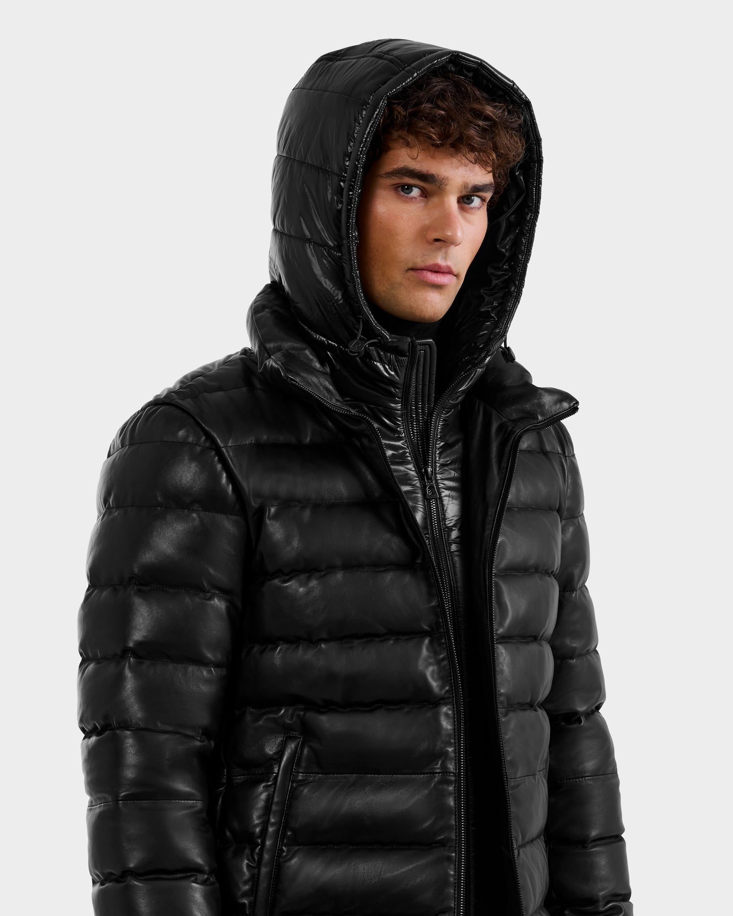 A young man with curly hair wears a shiny black Rudsak DRAVEN X - 6125686 men’s leather down jacket with the hood up, looking slightly to the side against a plain light background.