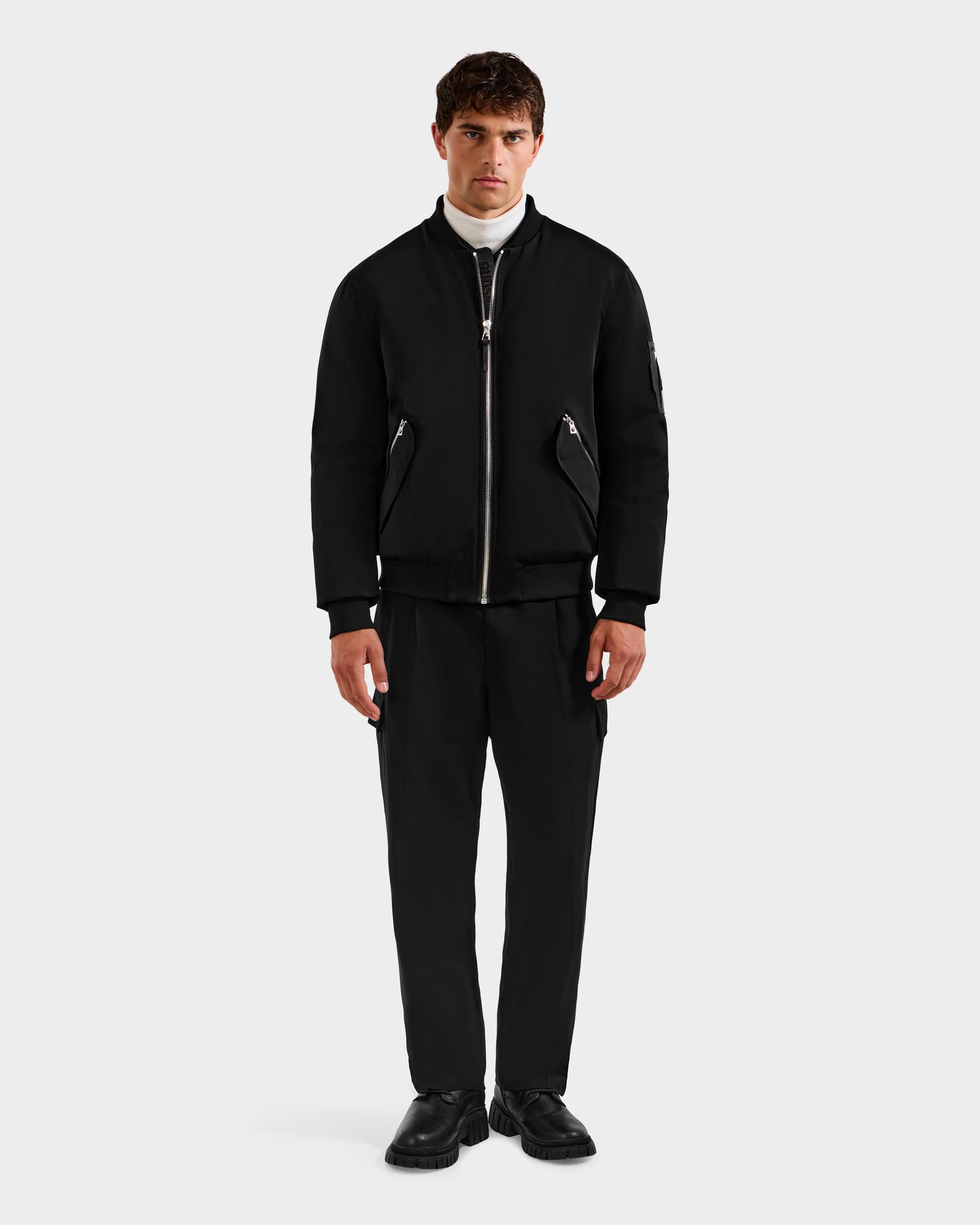 A man faces forward in the Rudsak DYLAN - 6125593 Black men’s aviator bomber jacket with removable faux fur collar, styled with black trousers, black shoes, and a white turtleneck against a plain light grey background.