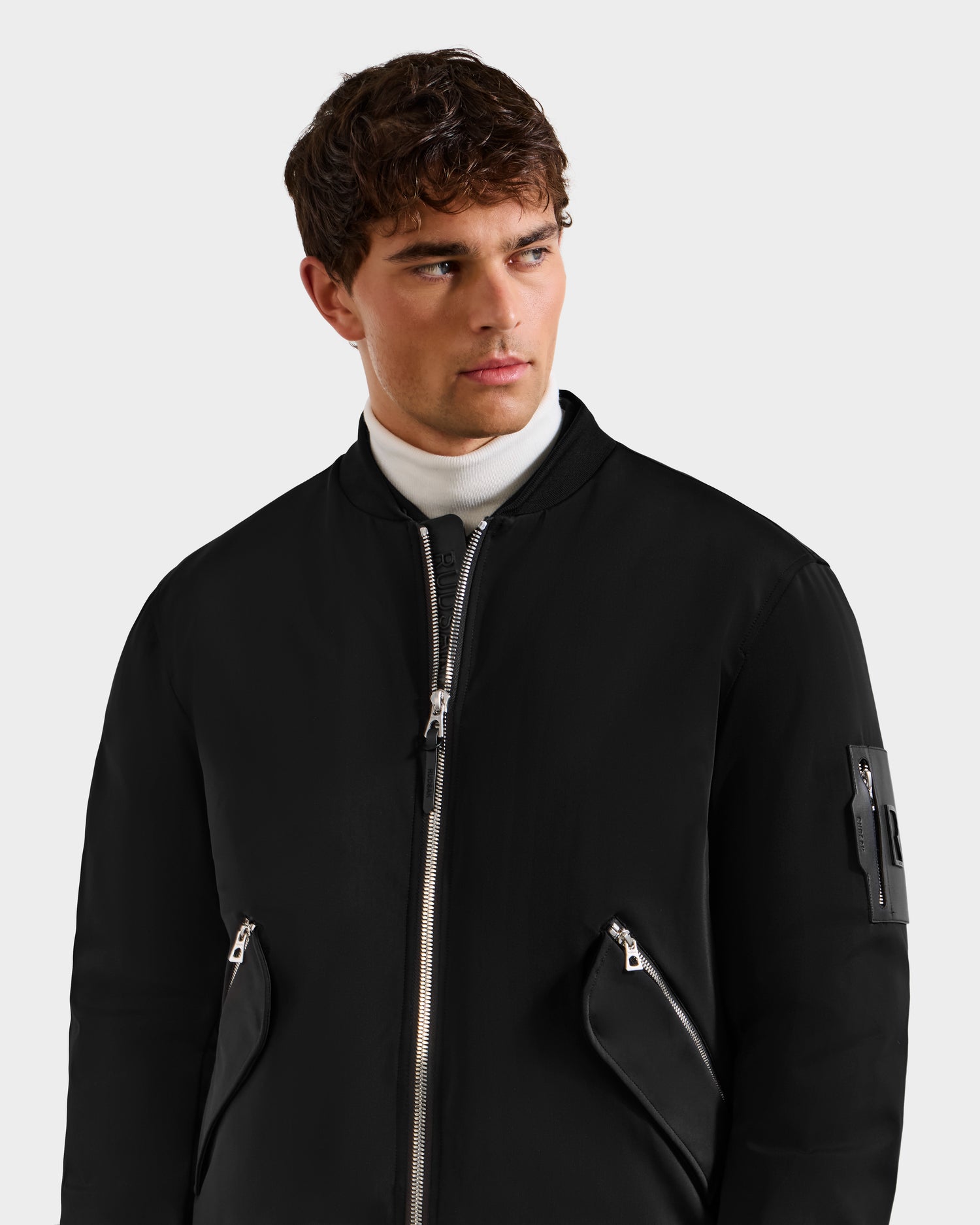 A young man in the Rudsak DYLAN - 6125593 black men's aviator bomber jacket with zippered pockets and a white turtleneck stands against a plain light background, gazing slightly left—a refined take on modern outerwear.