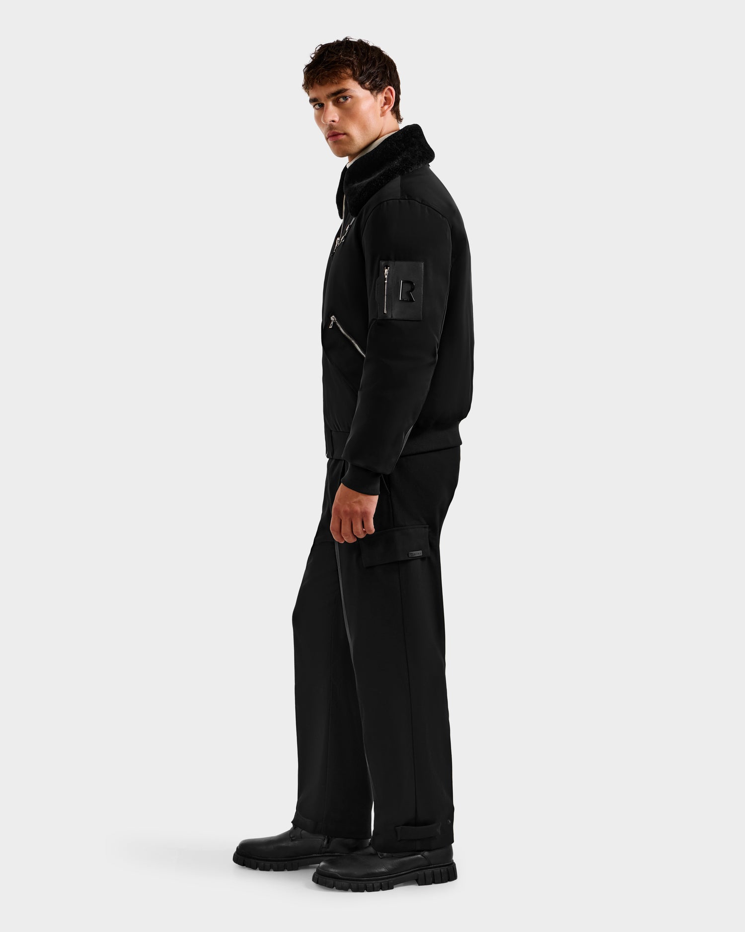 A man stands against a plain background wearing the Rudsak DYLAN - 6125593 Black aviator bomber jacket with a patch on the sleeve, paired with black pants and boots for a modern, timeless look.