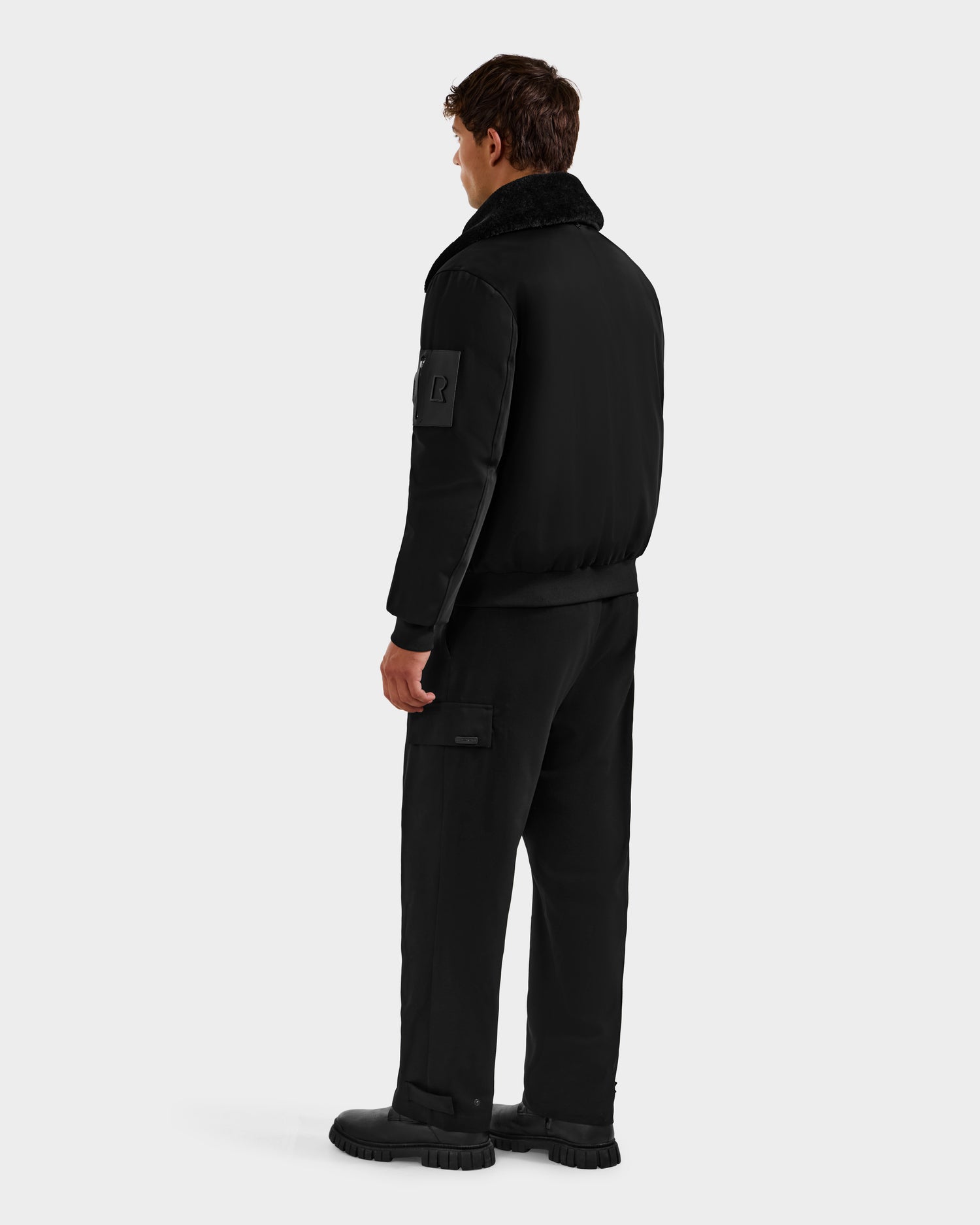 A person faces away, wearing Rudsak's DYLAN - 6125593 Black aviator bomber jacket with a removable faux fur collar, paired with black cargo pants and boots against a plain light gray background.