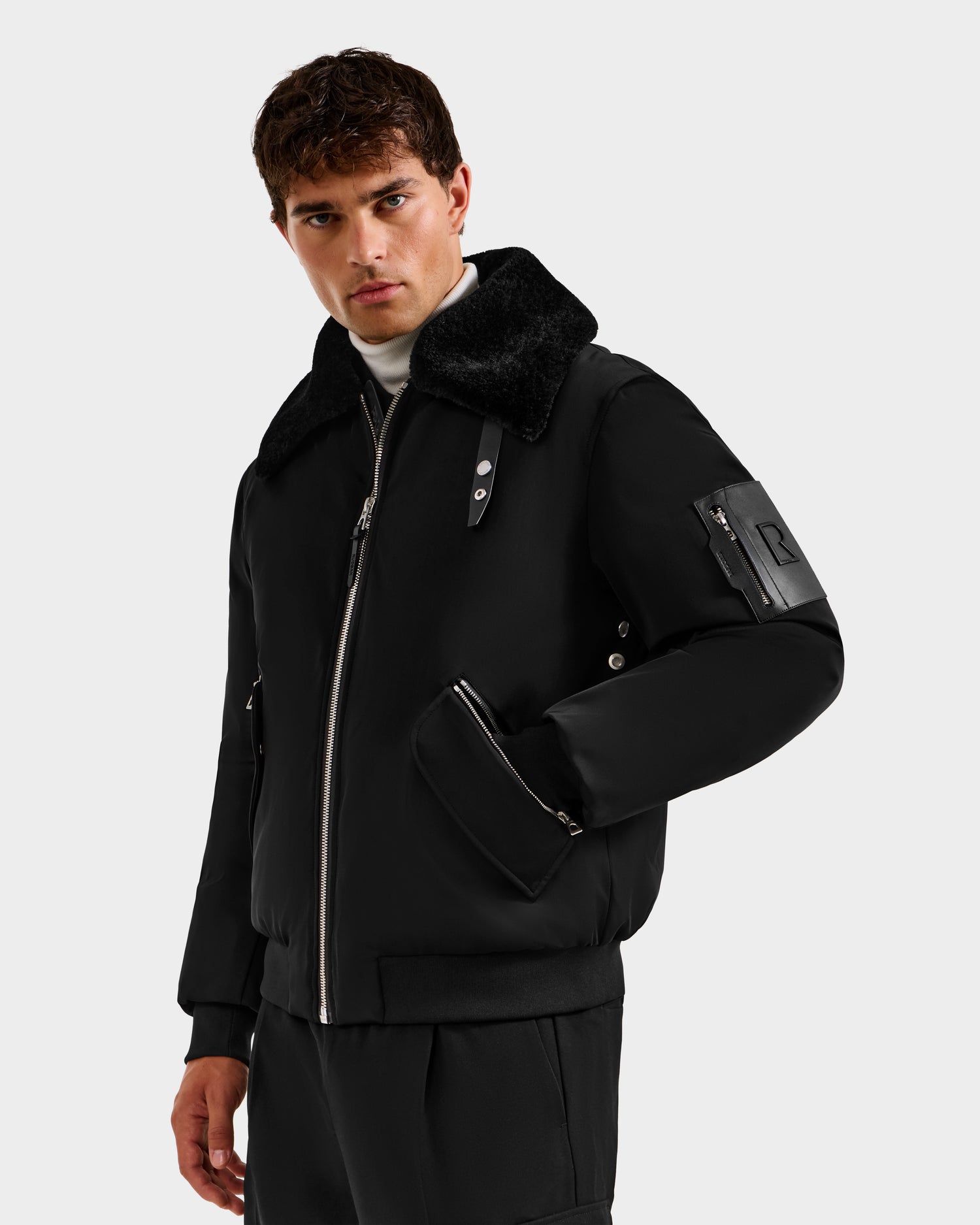 A man with short brown hair wears the Rudsak DYLAN - 6125593 Black men’s aviator bomber jacket with a removable faux fur collar, zipper details, and flap pockets, standing against a light gray background and looking confidently at the camera.