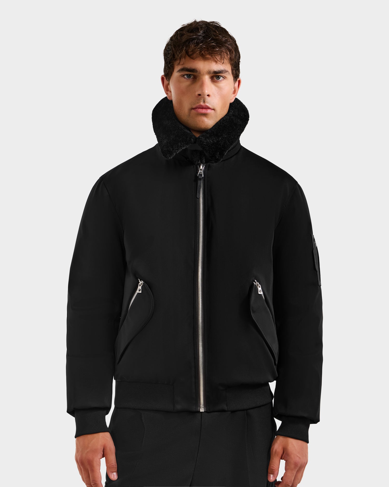 A young man faces forward in the Rudsak DYLAN - 6125593 Black men's aviator bomber jacket, featuring a zippered front and removable faux fur collar, paired with black pants against a light gray background for a modern look.