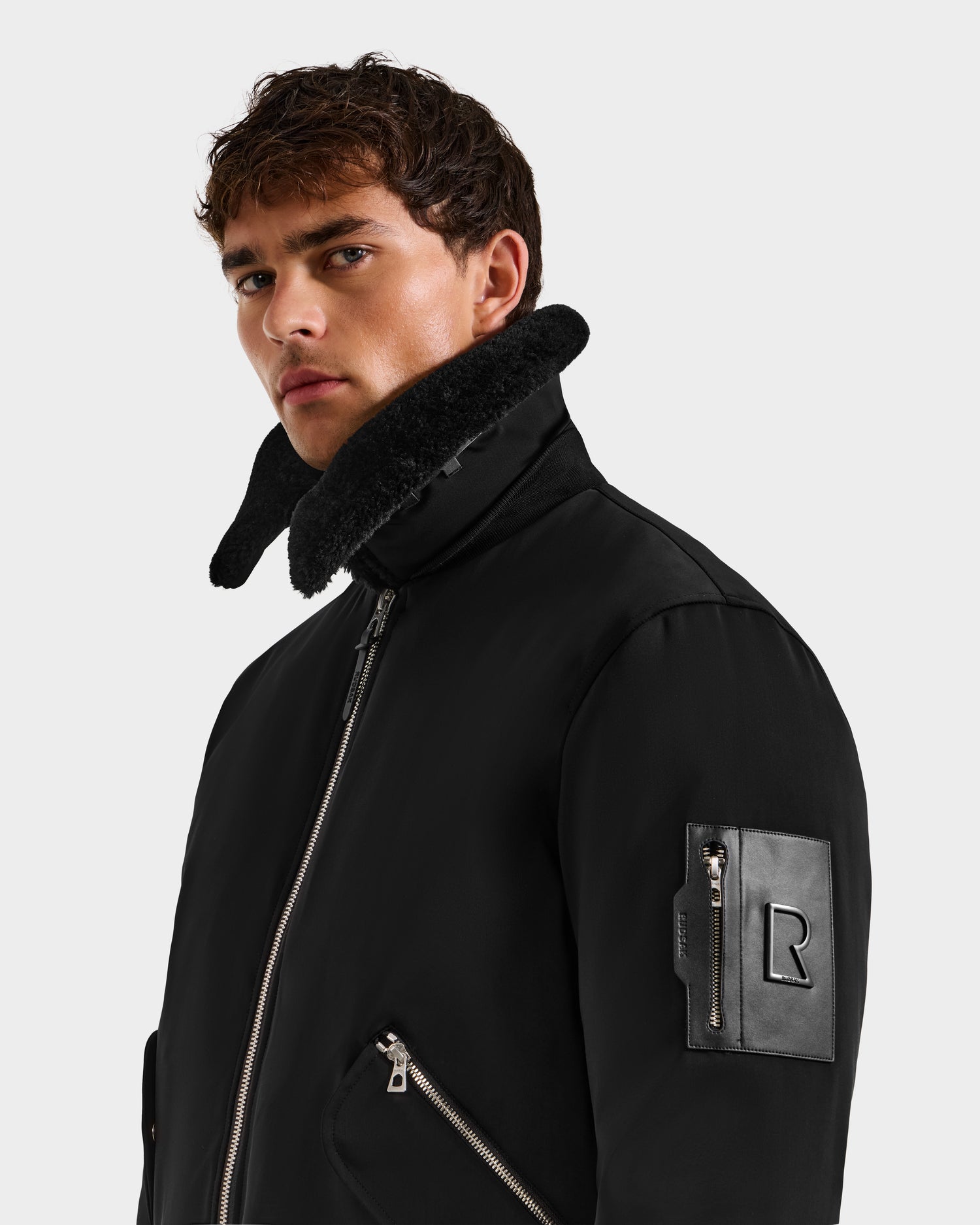 A young man with short brown hair wears the Rudsak DYLAN - 6125593 Black zip-up jacket with a high collar and leather sleeve patch, standing against a plain gray background and looking slightly to the side.