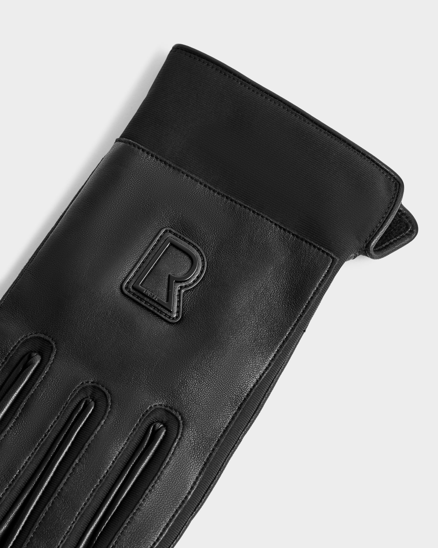 A close-up of Rudsak's EDDIE - 6425037 Black men's leather glove with decorative stitching and an embossed “R” on the back, photographed against a light gray background. These stylish gloves are ideal for staying warm in winter.