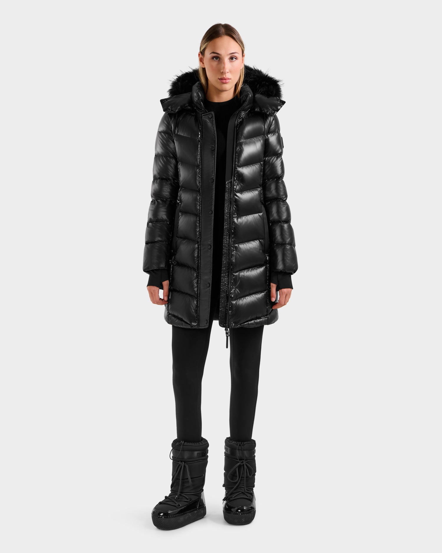 A woman faces forward against a plain light gray background, wearing the Rudsak ELISA S - L125572 Black shiny mid-length winter coat with a fur-trimmed hood, paired with black leggings and black winter boots.