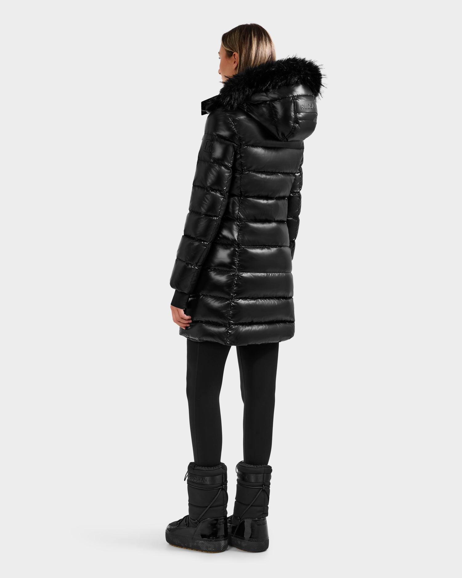 A person stands facing away, wearing the Rudsak ELISA S - L125572 Black women's down puffer with a fur-trimmed hood, paired with black leggings and winter boots against a plain light background.