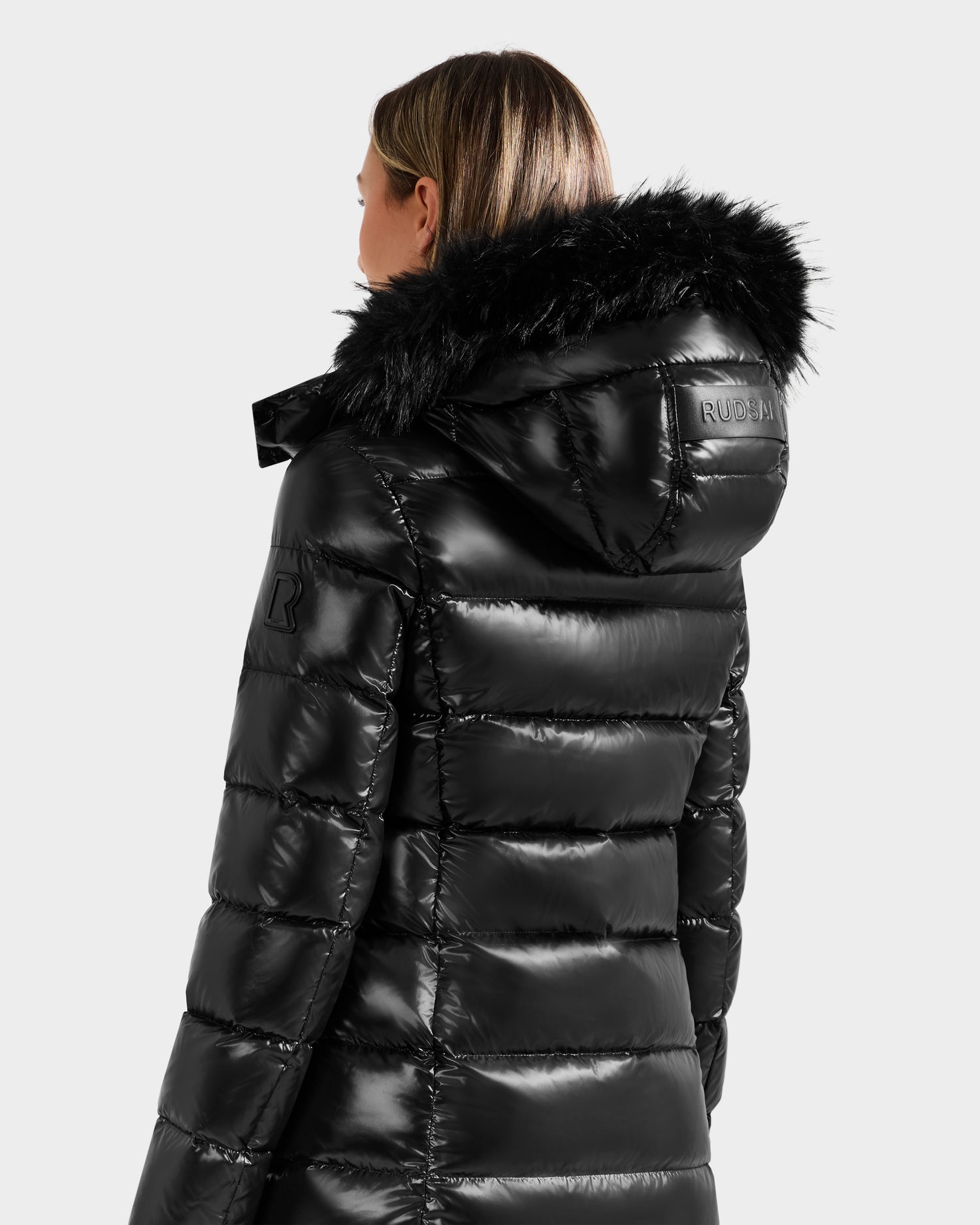 A woman is seen from behind wearing the Rudsak ELISA S - L125572 Black puffer jacket, a shiny black mid-length coat with a faux fur-trimmed hood, branded text on the back of the hood, and a patch on the sleeve.