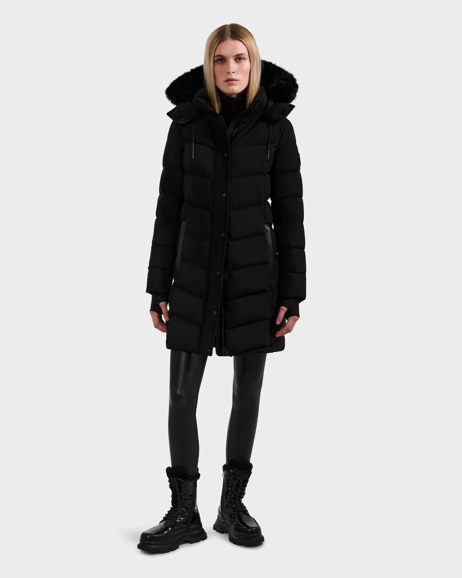 A woman faces forward wearing the Rudsak ELISA - L123516 Black long quilted down puffer with a fur-lined hood, paired with black leather pants, a turtleneck, and lace-up boots against a light gray background.