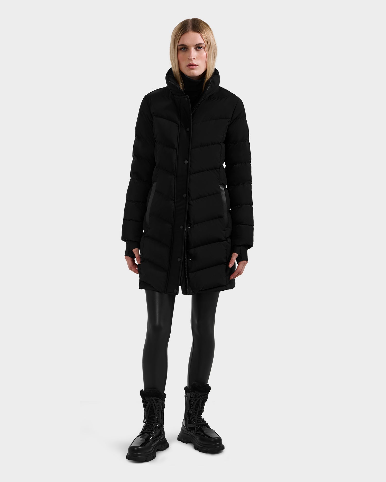 A person wears the Rudsak ELISA - L123516 Black women's quilted down puffer coat, paired with black leggings and black lace-up boots, standing forward against a plain light background.