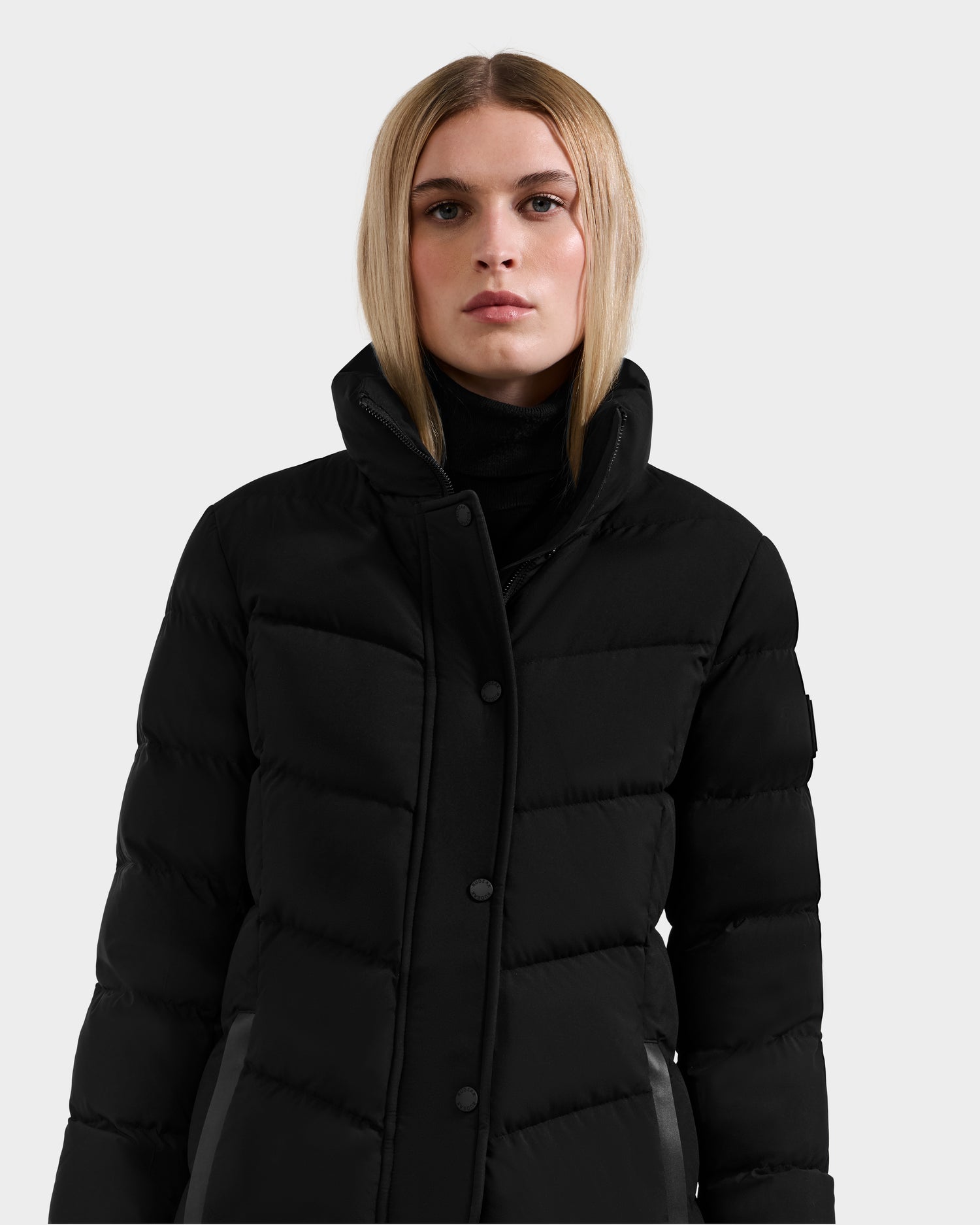 A woman with straight blonde hair wears the Rudsak ELISA - L123516 Black women’s quilted down puffer made with RDS-certified down, standing against a plain light gray background.