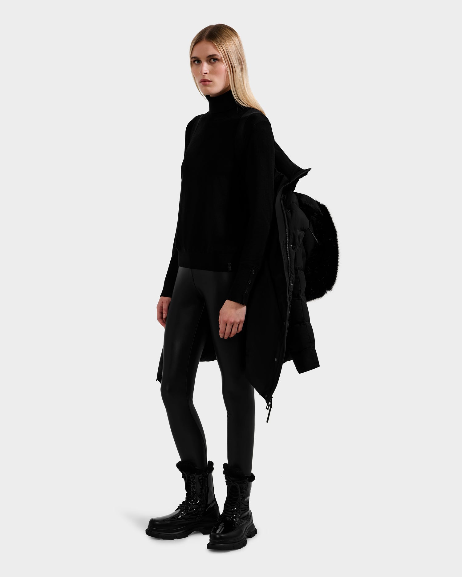A woman with long blonde hair wears Rudsak's ELISA - L123516 Black sustainable winter coat over one shoulder, paired with a black turtleneck, shiny leggings, and combat boots against a plain light gray background.