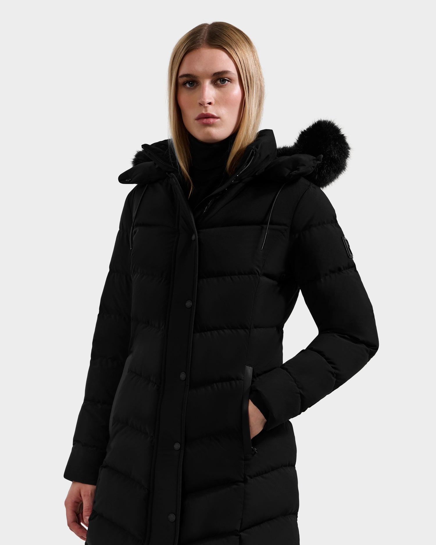 A woman wears the Rudsak ELISA - L123516 Black women's long quilted down puffer coat with a black faux fur-trimmed hood, standing with one hand in her pocket and looking ahead against a plain light background.