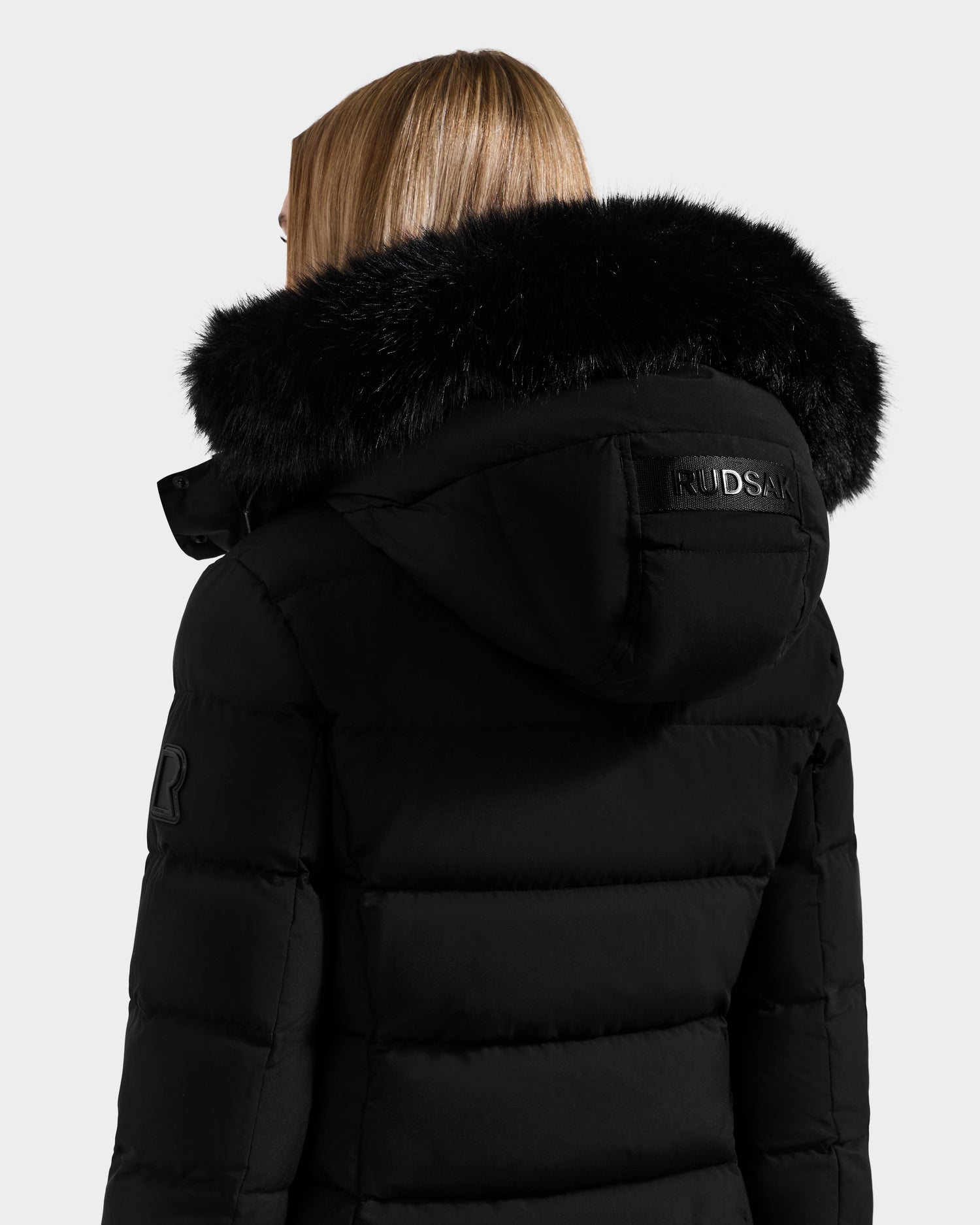 A person with straight, light brown hair wears the Rudsak ELISA - L123516 Black women’s quilted down puffer jacket with a large fur-lined hood. The sustainable coat’s Rudsak logo patch is visible against the plain white background.