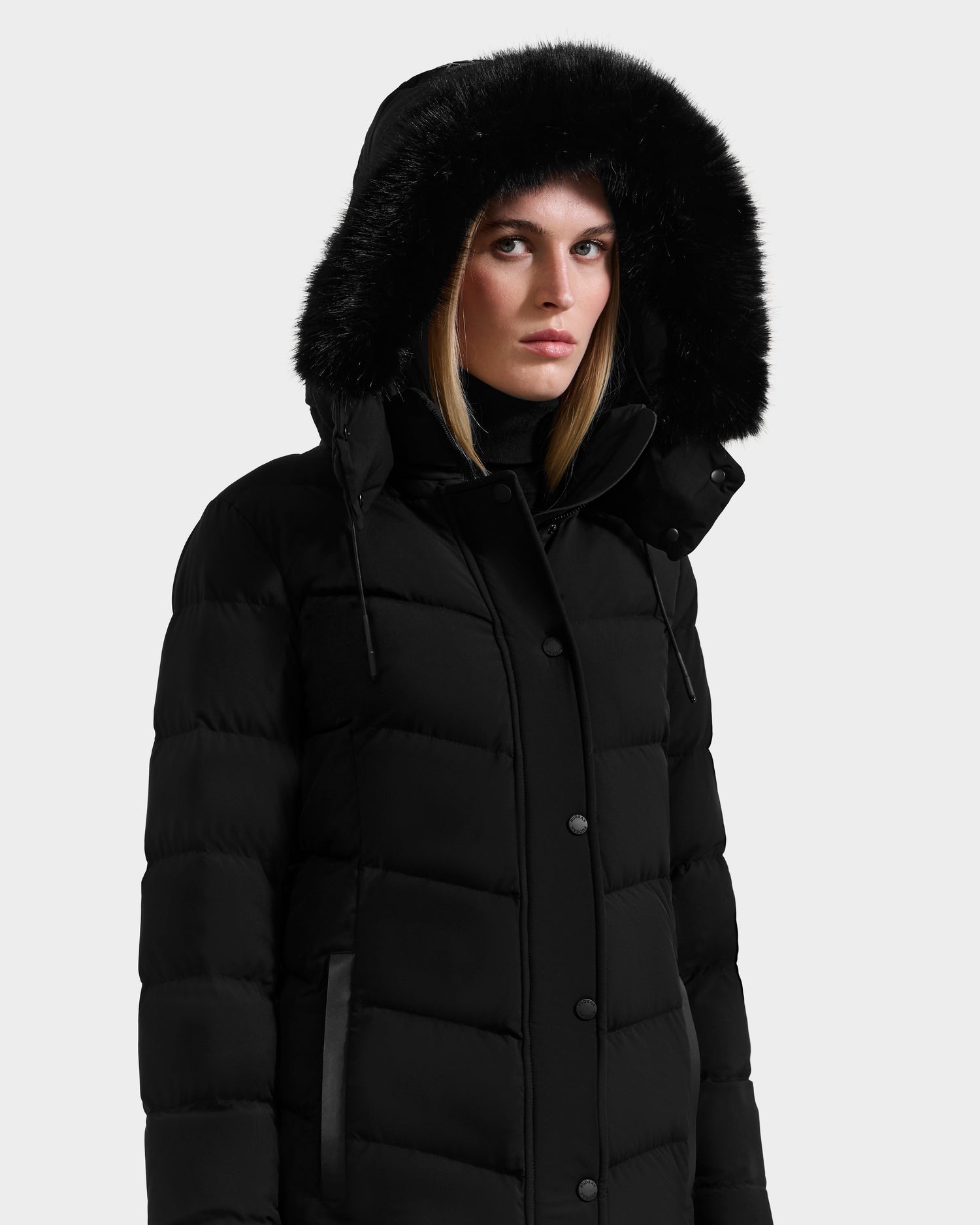 A woman wearing the Rudsak ELISA - L123516 Black quilted down puffer with furry trim stands against a plain light background, looking directly at the camera with a neutral expression.