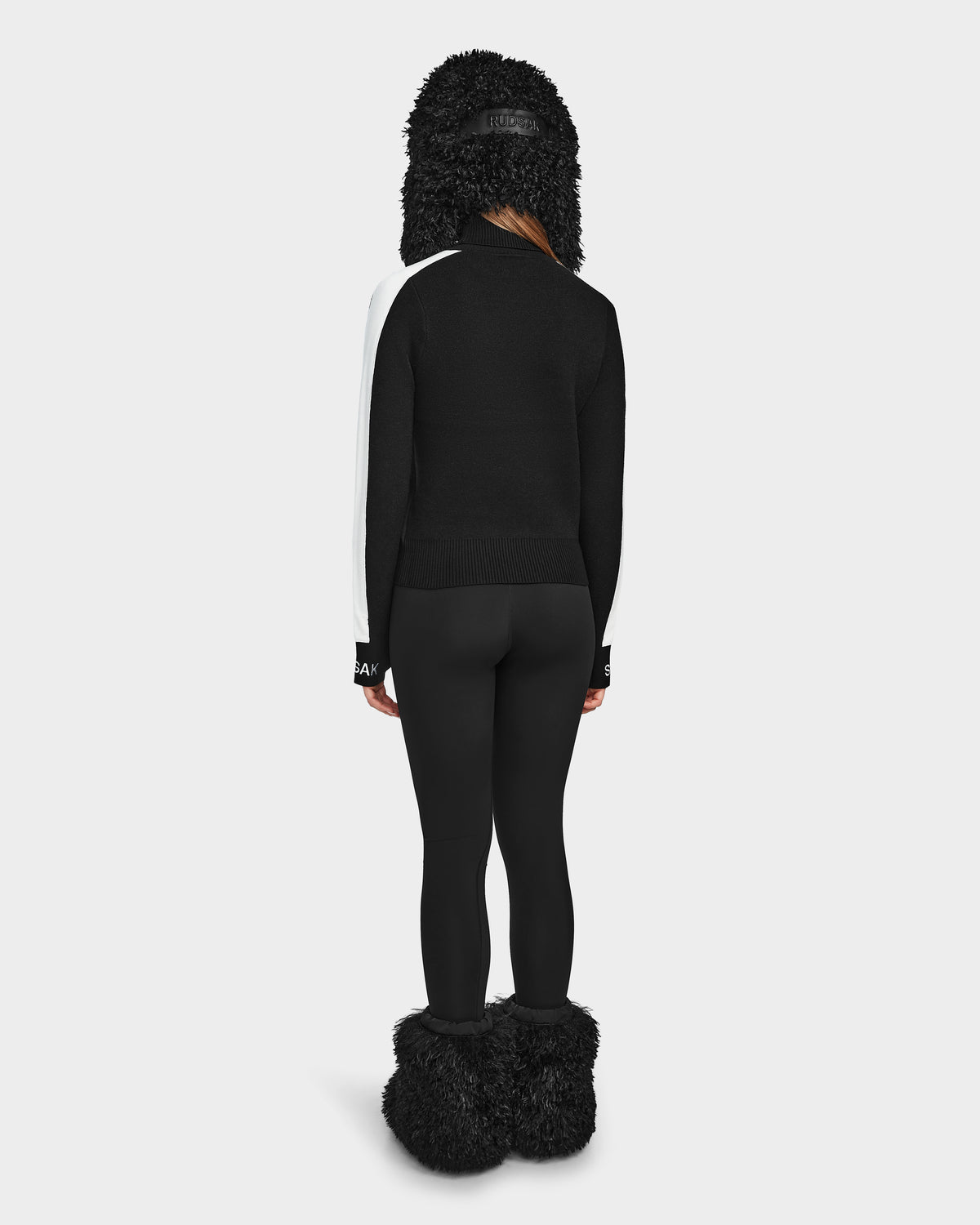 A woman models the Rudsak ELIZABETH - 8524623 Black winter jacket, paired with a womens ski turtleneck, black leggings, furry boots, and a black furry ear-flap hat, all set against a plain white background.