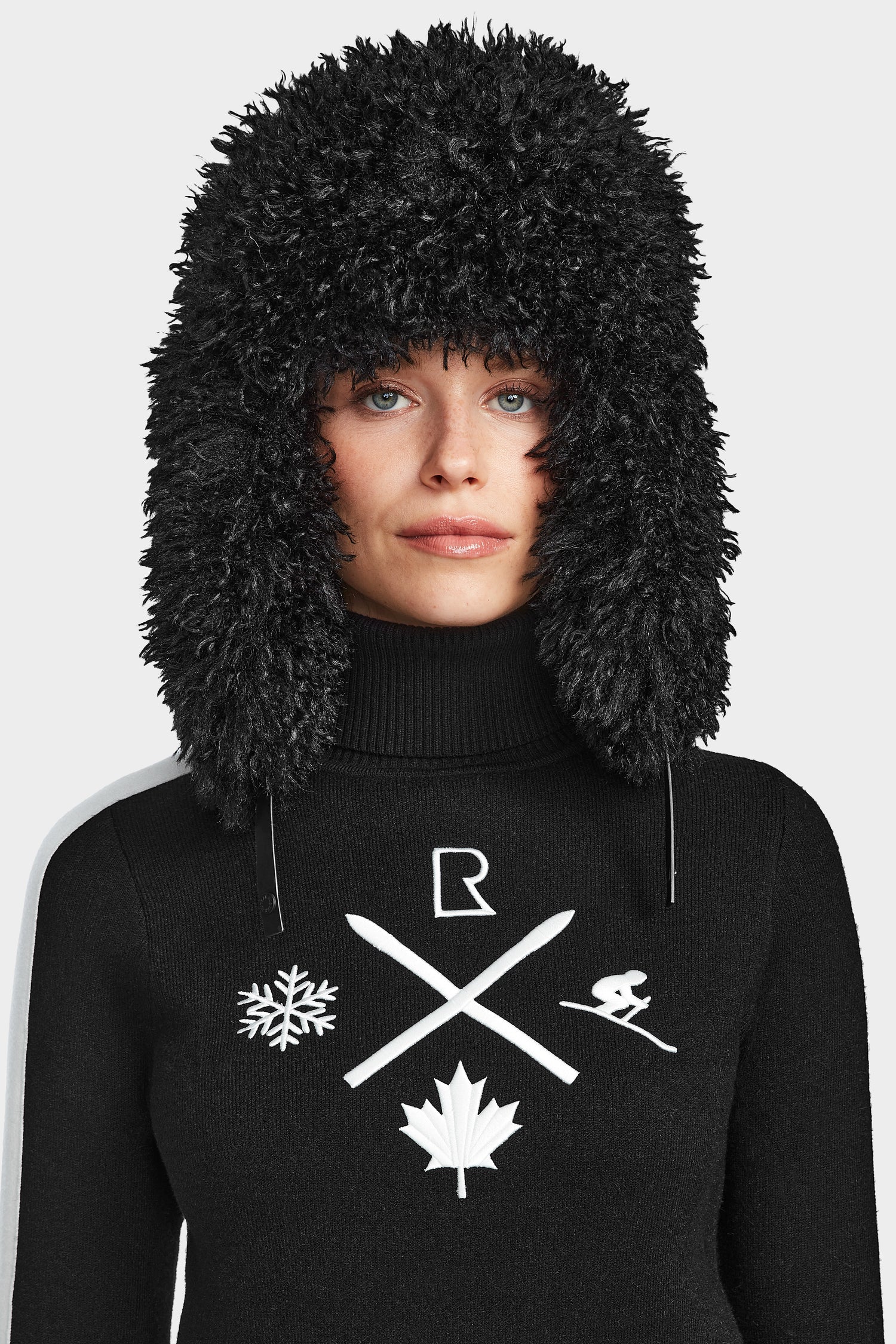 A woman wears the Rudsak ELIZABETH - 8524623 Black, a large, fluffy black winter hat, with a womens ski turtleneck sweater featuring white snowflake, ski poles, maple leaf, and skier graphics—ideal for winter or après-ski style.