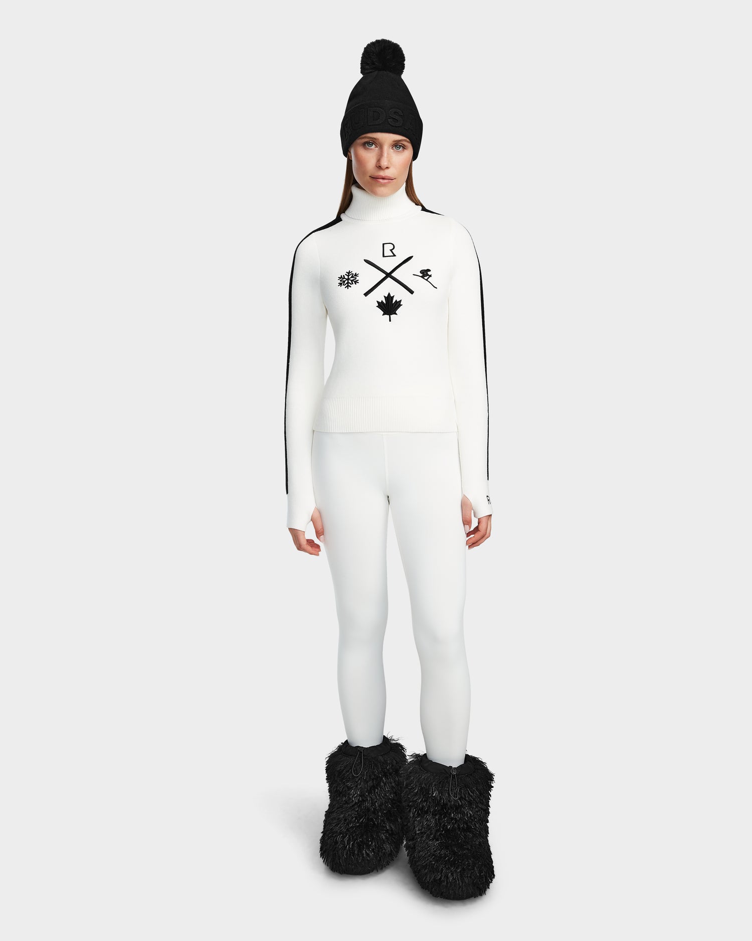 A woman faces forward in a chic après-ski look, wearing the Rudsak ELIZABETH - 8524623 Optic White ski turtleneck with black symbols, matching leggings, a black pom-pom beanie, and large black furry boots against a light plain background.