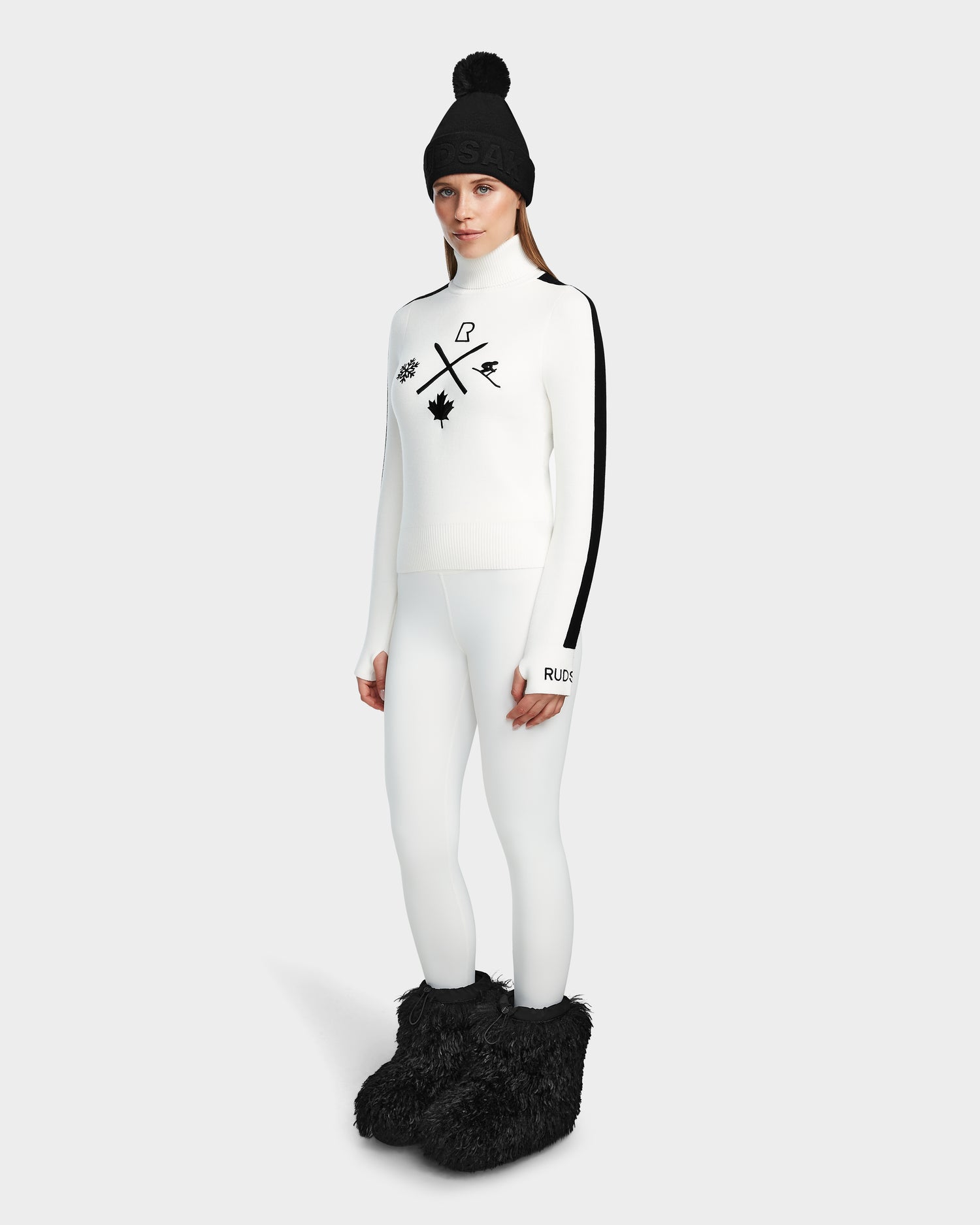 A woman faces forward in the Rudsak ELIZABETH - 8524623 Optic White ski turtleneck, paired with white leggings, a black beanie, and black furry boots—delivering chic winter style against a plain light gray background.