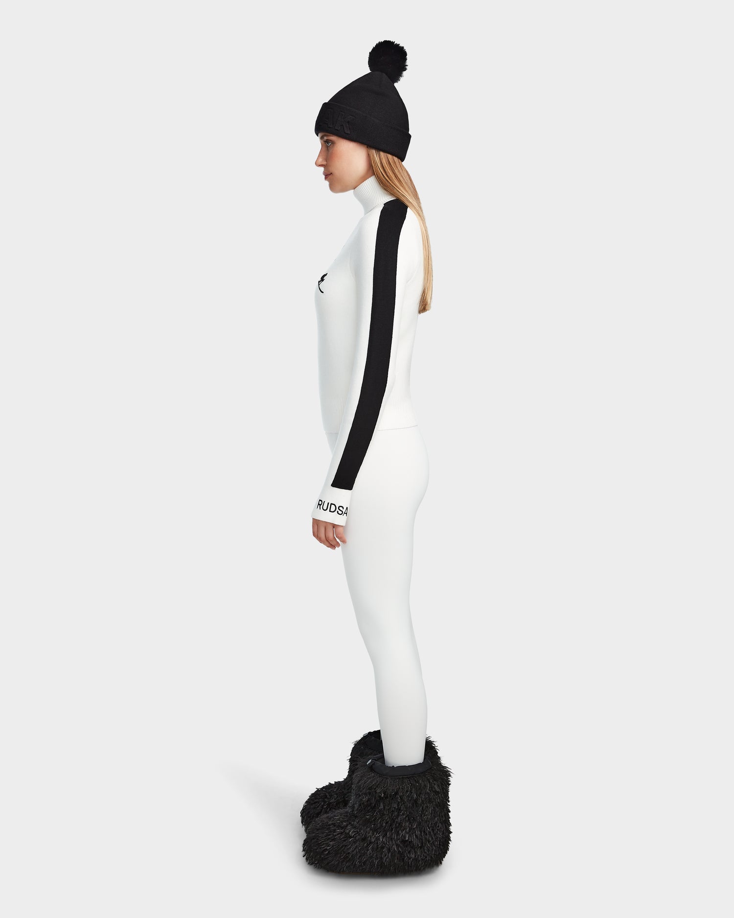 A woman in the Rudsak ELIZABETH - 8524623 Optic White ski turtleneck with black sleeves, a black pom-pom beanie, and furry black boots stands in profile against a light gray backdrop—ideal for a chic winter wardrobe.