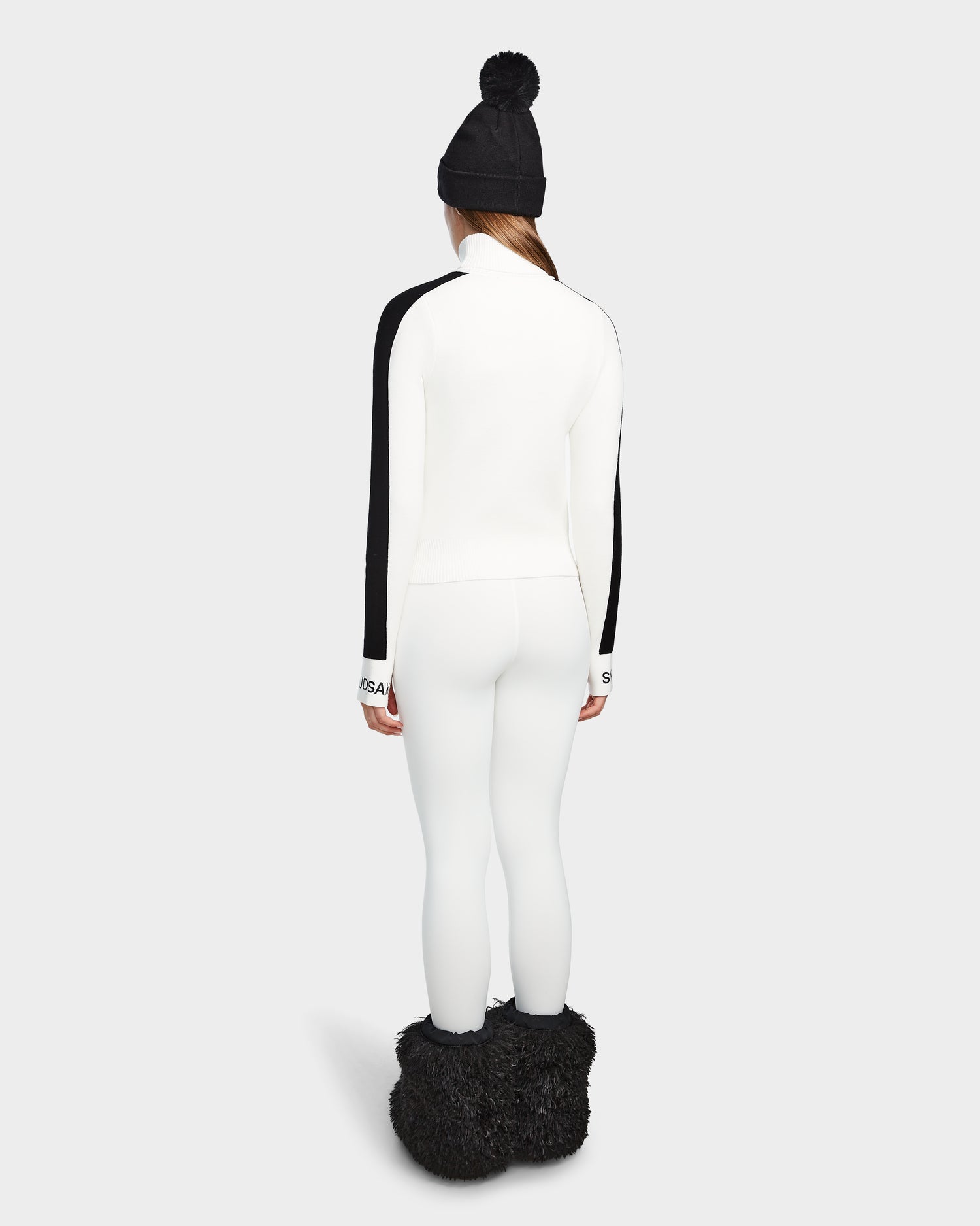 A person stands facing away, styled in Rudsak’s ELIZABETH - 8524623 Optic White turtleneck, a black beanie, white leggings, and black furry boots—an effortlessly chic winter look set against a simple light background.