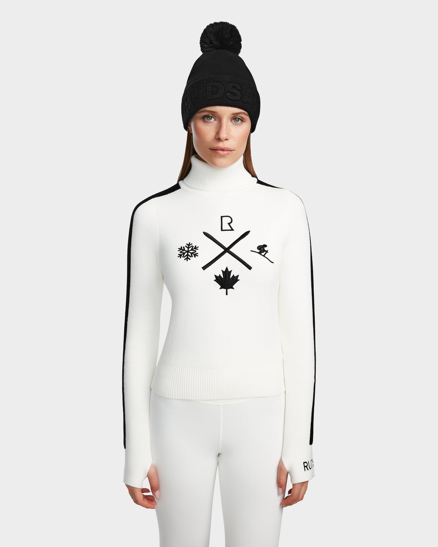 A woman in a black beanie and ski turtleneck with black winter symbols pairs it with Rudsaks ELIZABETH - 8524623 Optic White pants, creating a chic après-ski look against a light gray background.