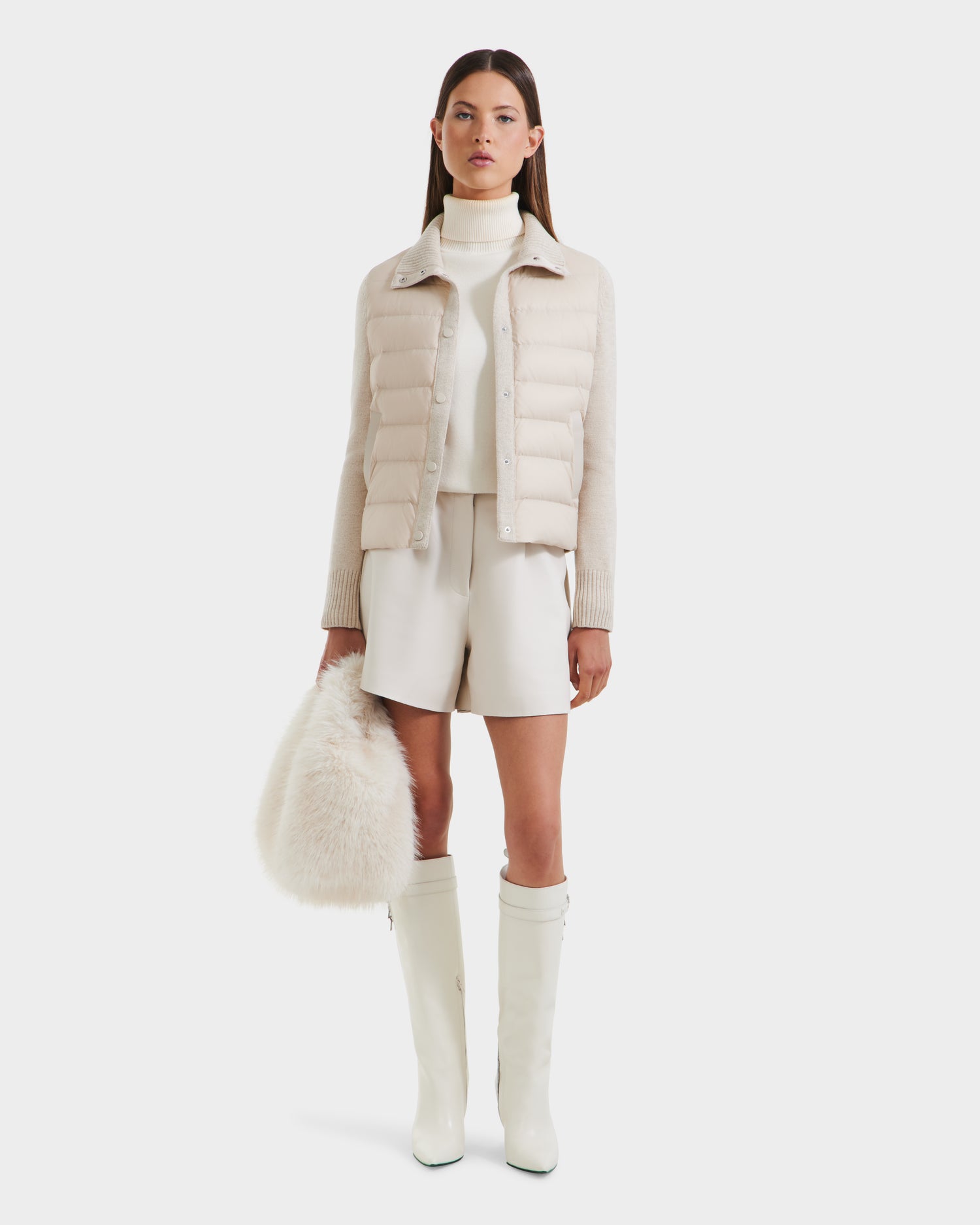 A woman stands facing forward against a plain background, dressed in Rudsak’s ELLIE - 8125635 Vanilla hybrid down jacket—perfect for transitional layering with a white turtleneck, shorts, knee-high boots, and a fluffy white handbag.