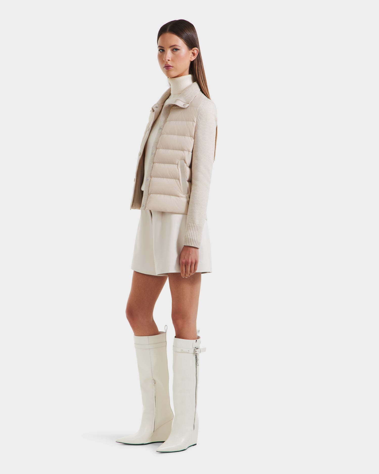 A woman stands sideways against a white background, wearing the Rudsak ELLIE - 8125635 Vanilla light beige quilted down jacket over a cream turtleneck, short cream skirt, and knee-high white zippered boots.
