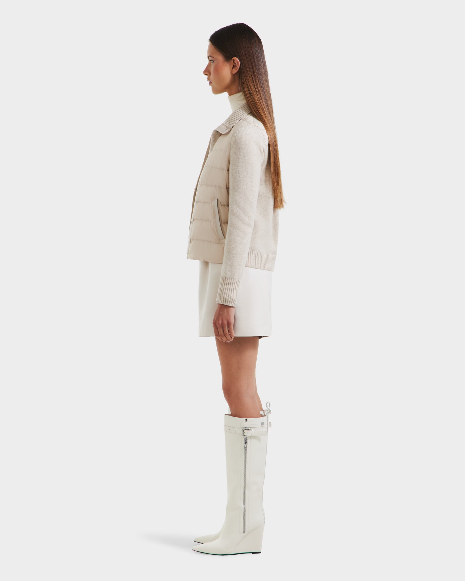 A woman stands in profile against a plain white background wearing the Rudsak ELLIE - 8125635 Vanilla quilted down jacket, paired with a white mini skirt and knee-high white zippered boots—an ideal look for transitional layering.