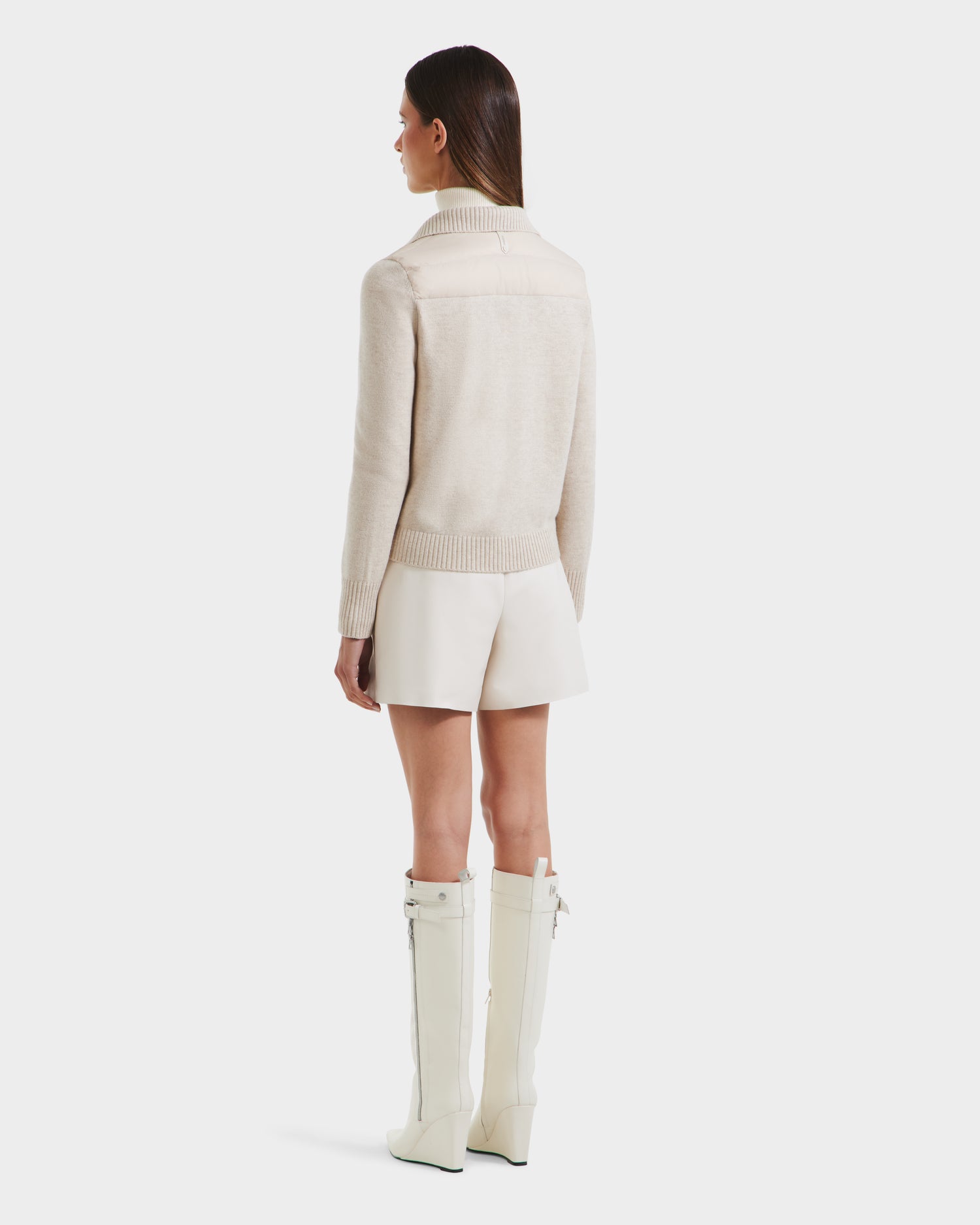 A woman faces away against a white background, wearing the Rudsak ELLIE - 8125635 Vanilla quilted down jacket, paired with white shorts and tall white heeled boots.