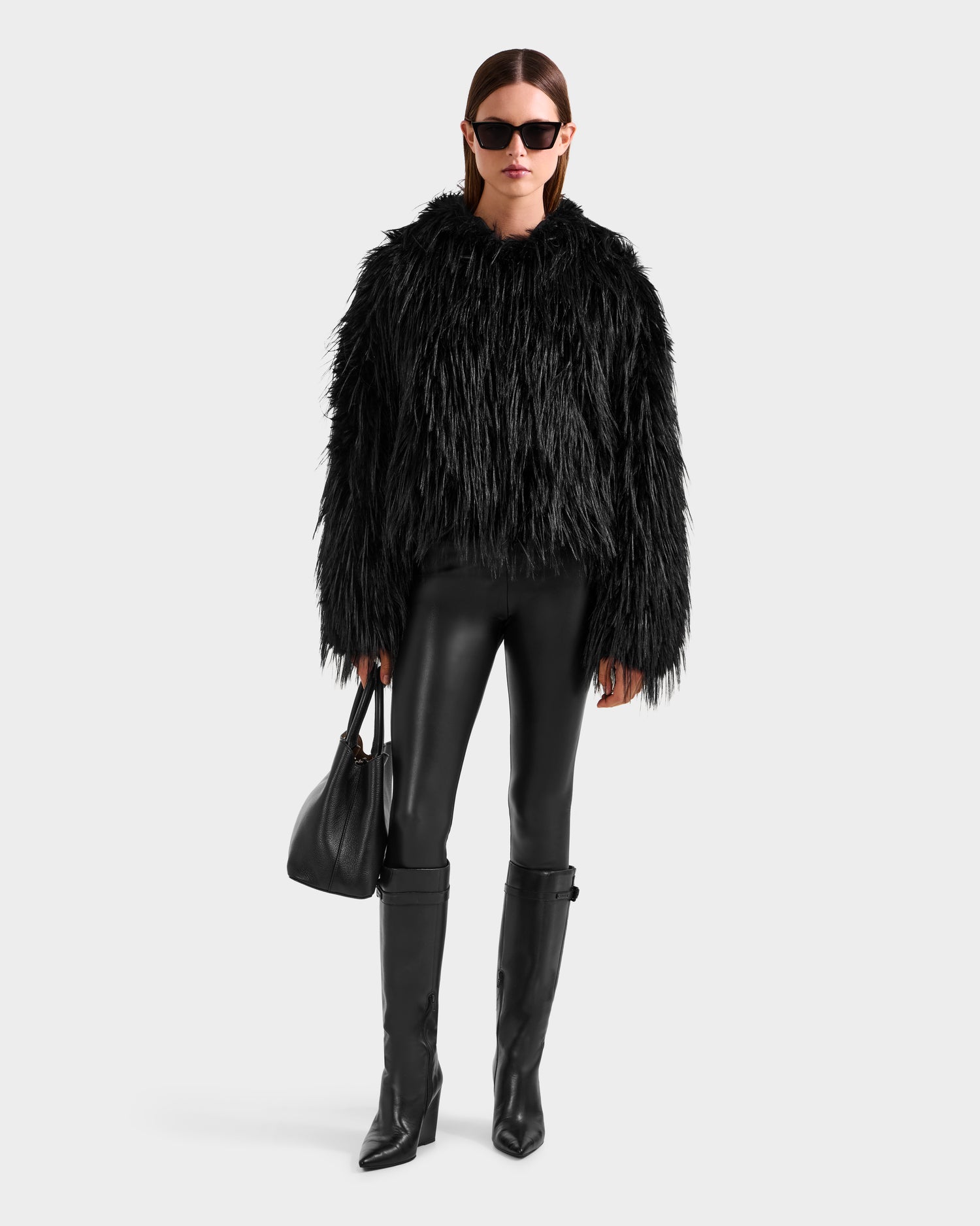 Dressed in the Rudsak ELLISON - 8525678 Black plush faux fur hoodie, black leather pants, and knee-high black boots, a person with sunglasses and a black handbag showcases an après-ski look against a light gray background.