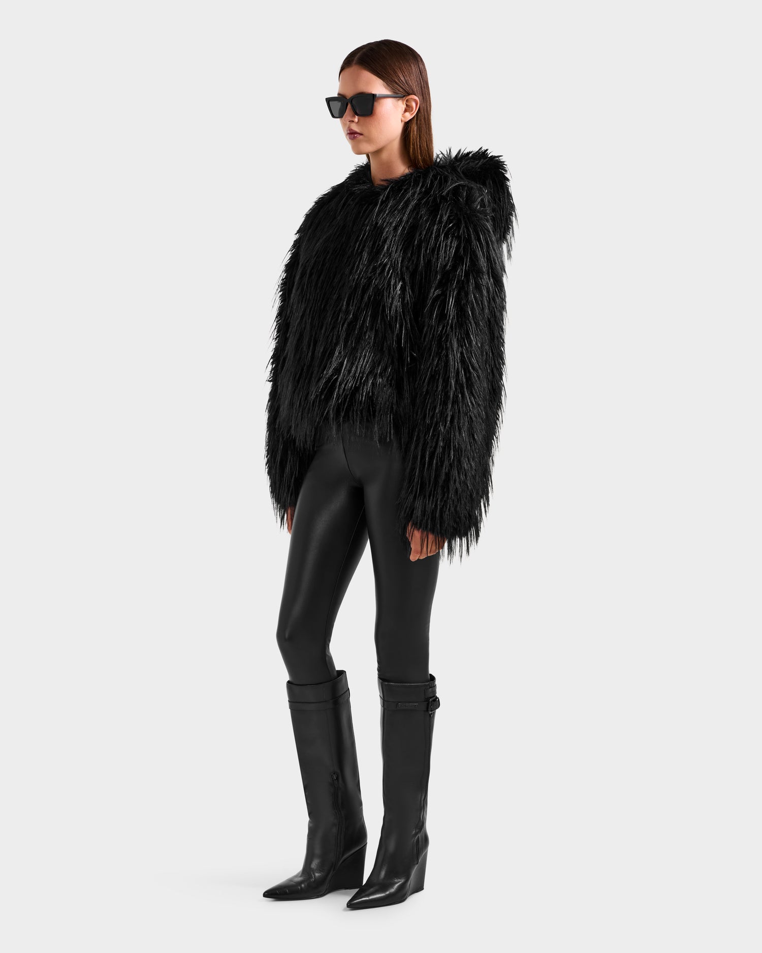 A person wearing Rudsak’s ELLISON - 8525678 Black shaggy faux fur hoodie with an oversized hood stands in shiny black leggings and knee-high boots against a light gray background.