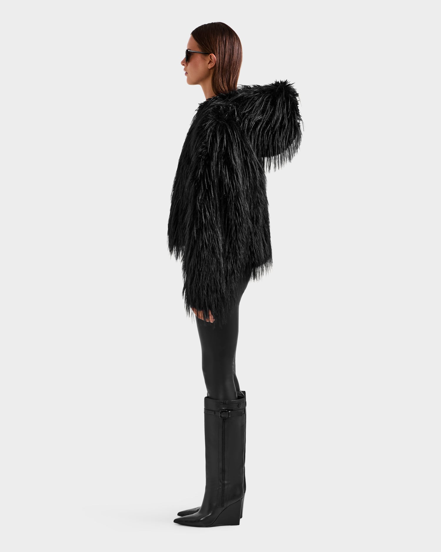A person in profile wears Rudsak's ELLISON - 8525678 Black sunglasses, a plush faux fur hoodie with an oversized hood, black leggings, and knee-high black wedge boots against a plain light gray background.