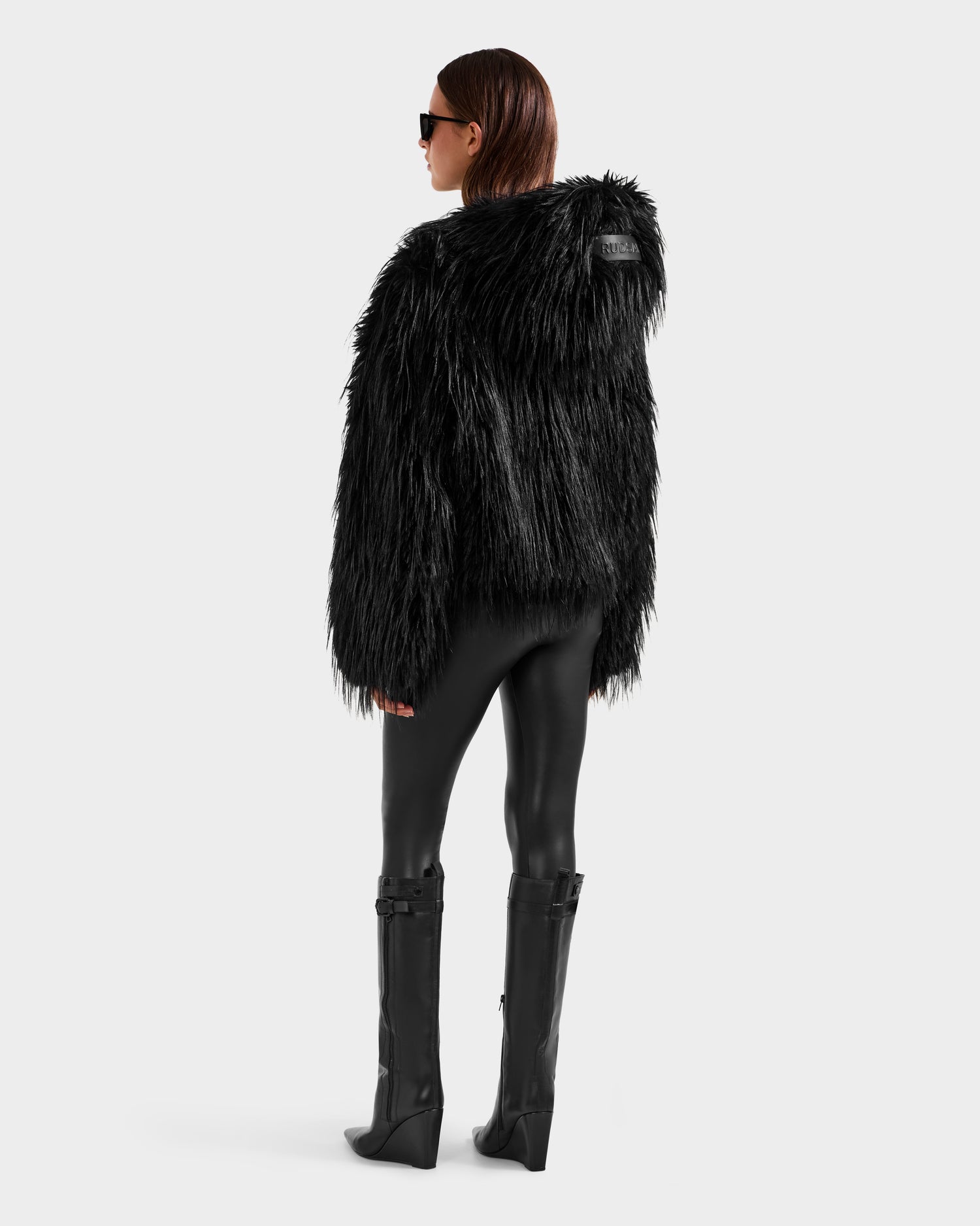 A person in the Rudsak ELLISON - 8525678 Black plush faux fur hoodie with an oversized hood, black shiny leggings, tall black boots, and sunglasses stands against a plain background—effortlessly channeling après-ski style.