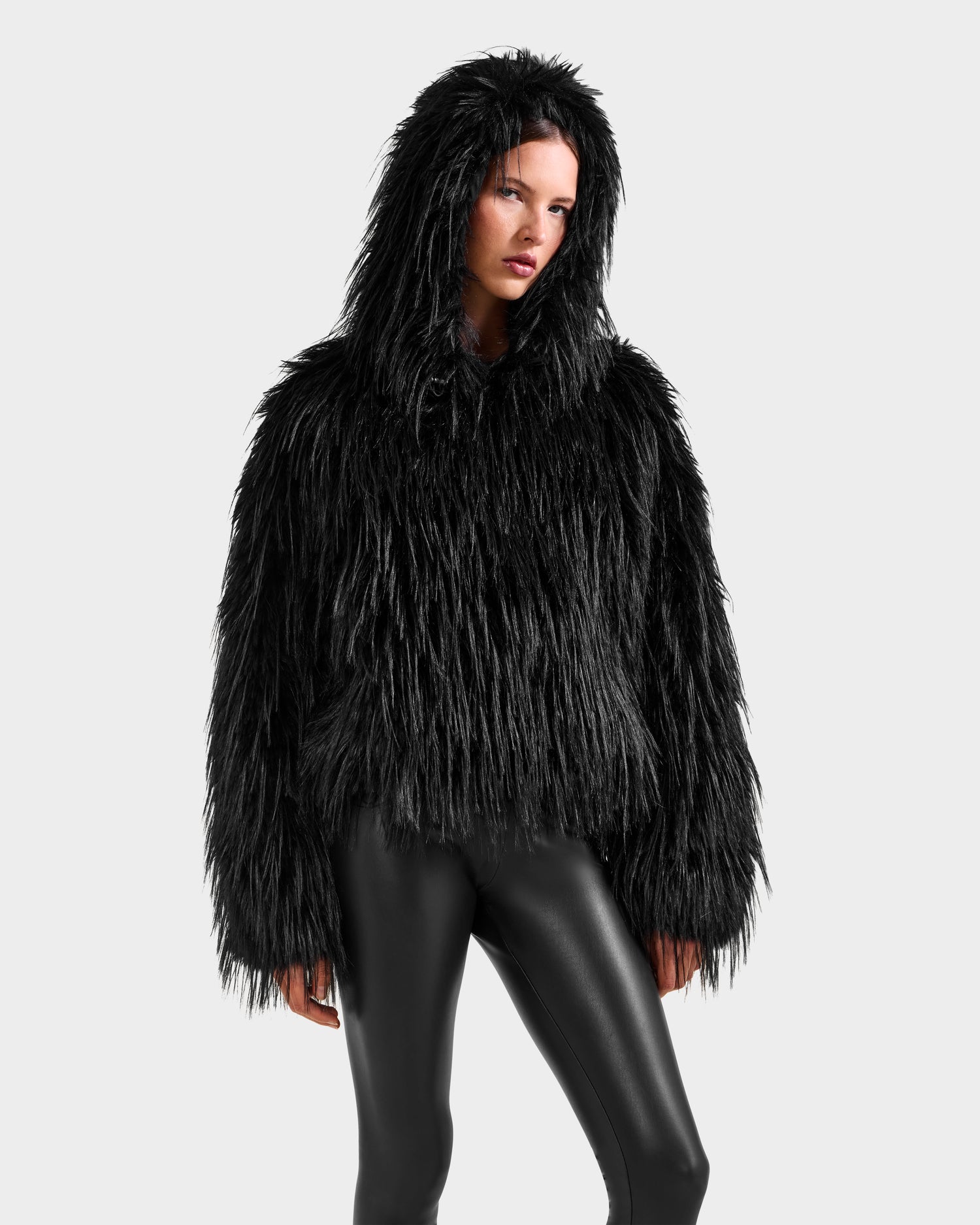 A person showcases the Rudsak ELLISON - 8525678 Black plush faux fur hoodie with an oversized hood and shiny black leggings against a light gray backdrop, embodying an effortless après-ski vibe.