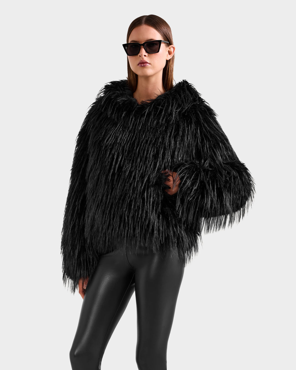 A woman models the Rudsak ELLISON - 8525678 Black, featuring black sunglasses, a plush black faux fur hoodie with an oversized hood, and shiny leggings, posing stylishly against a light gray background.