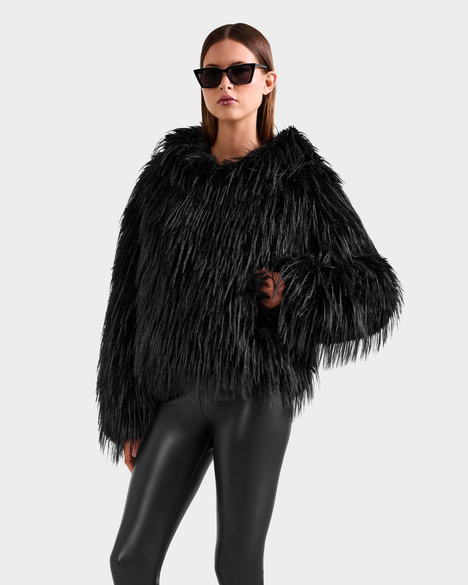 A woman models the Rudsak ELLISON - 8525678 Black, featuring black sunglasses, a plush black faux fur hoodie with an oversized hood, and shiny leggings, posing stylishly against a light gray background.