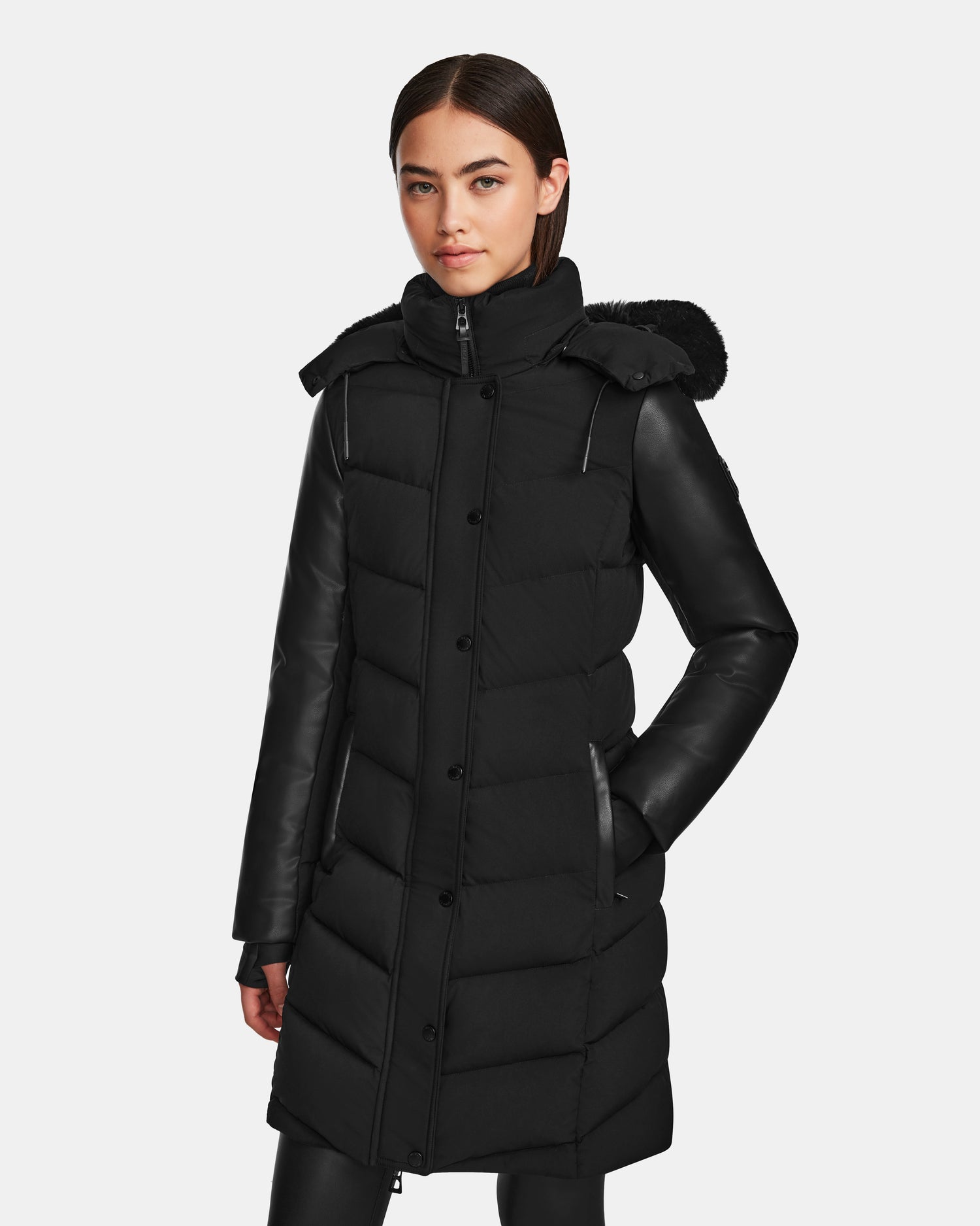 A woman with straight dark hair models the Rudsak ELSA - L123535 Black womens quilted down puffer coat with leather sleeves and a hood, standing against a plain light background with one hand in her pocket.