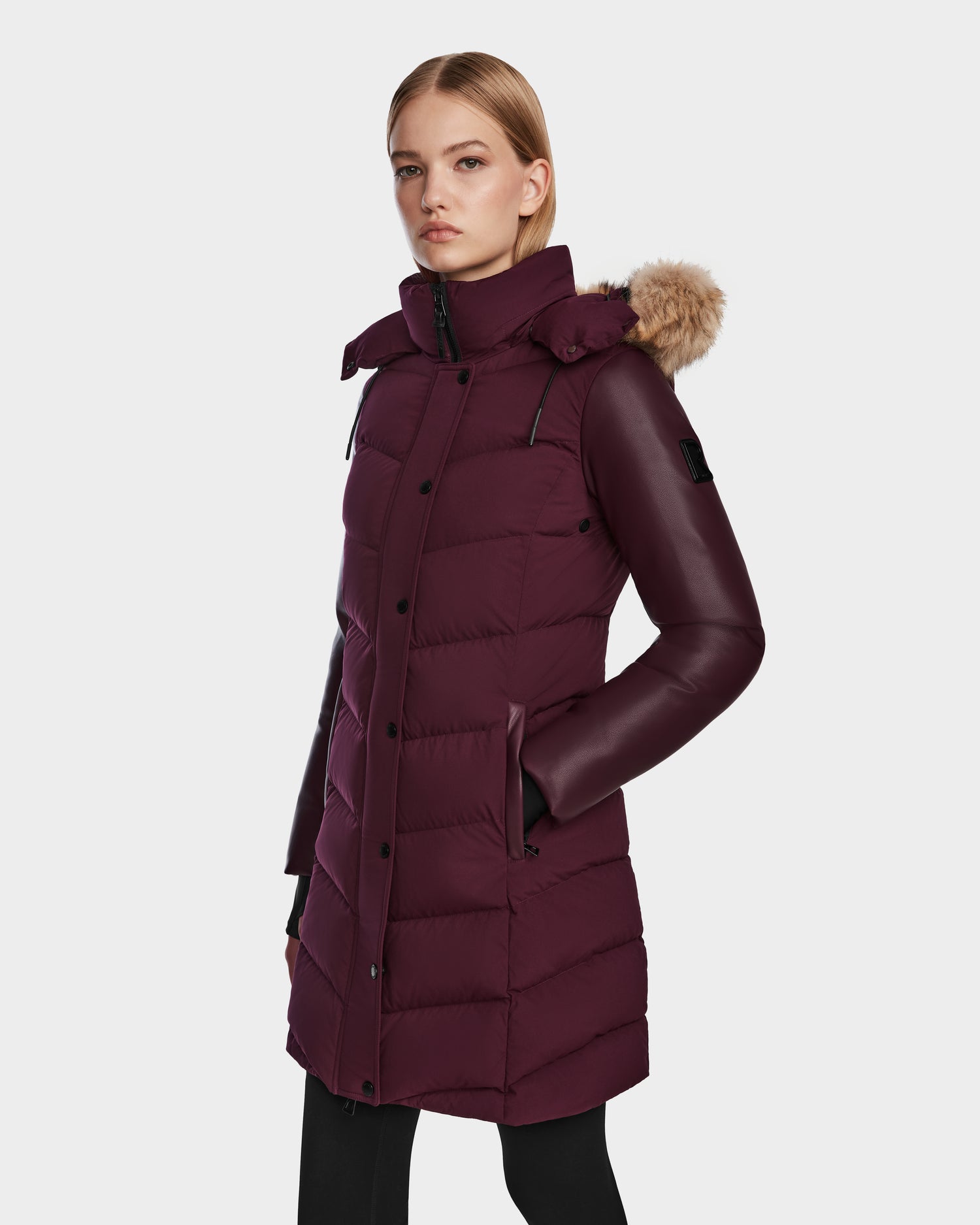 A woman stands facing forward against a plain white background, wearing the Rudsak ELSA - L123535 Wine, a long quilted down puffer with a fur-trimmed hood.