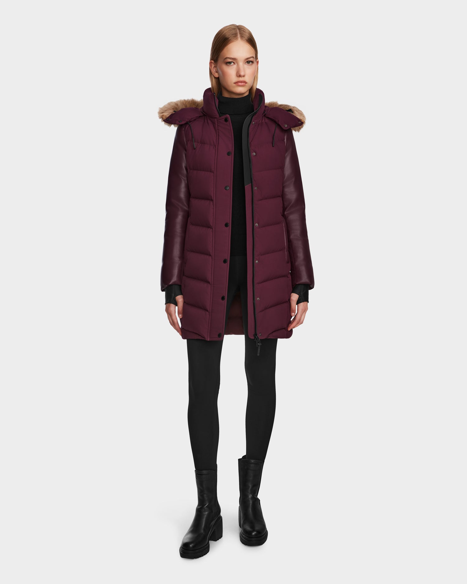 A woman wears the Rudsak ELSA - L123535 Wine quilted down puffer with a fur-trimmed hood, styled with black leggings, a turtleneck, and ankle boots against a plain white background.
